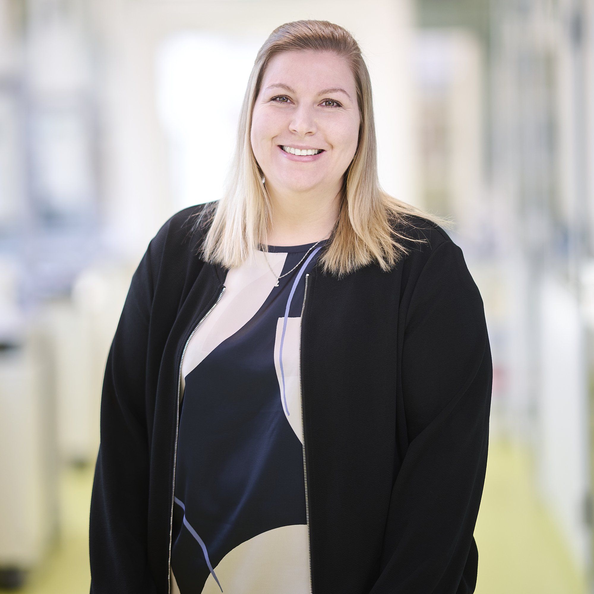 A woman with shoulder-length blonde hair smiling, wearing a black jacket over a patterned blouse, standing in a well-lit indoor space with blurred background.