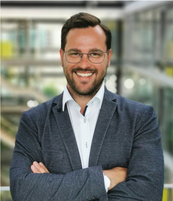 Smiling man with glasses and a beard in a gray blazer and white shirt, standing with arms crossed indoors.