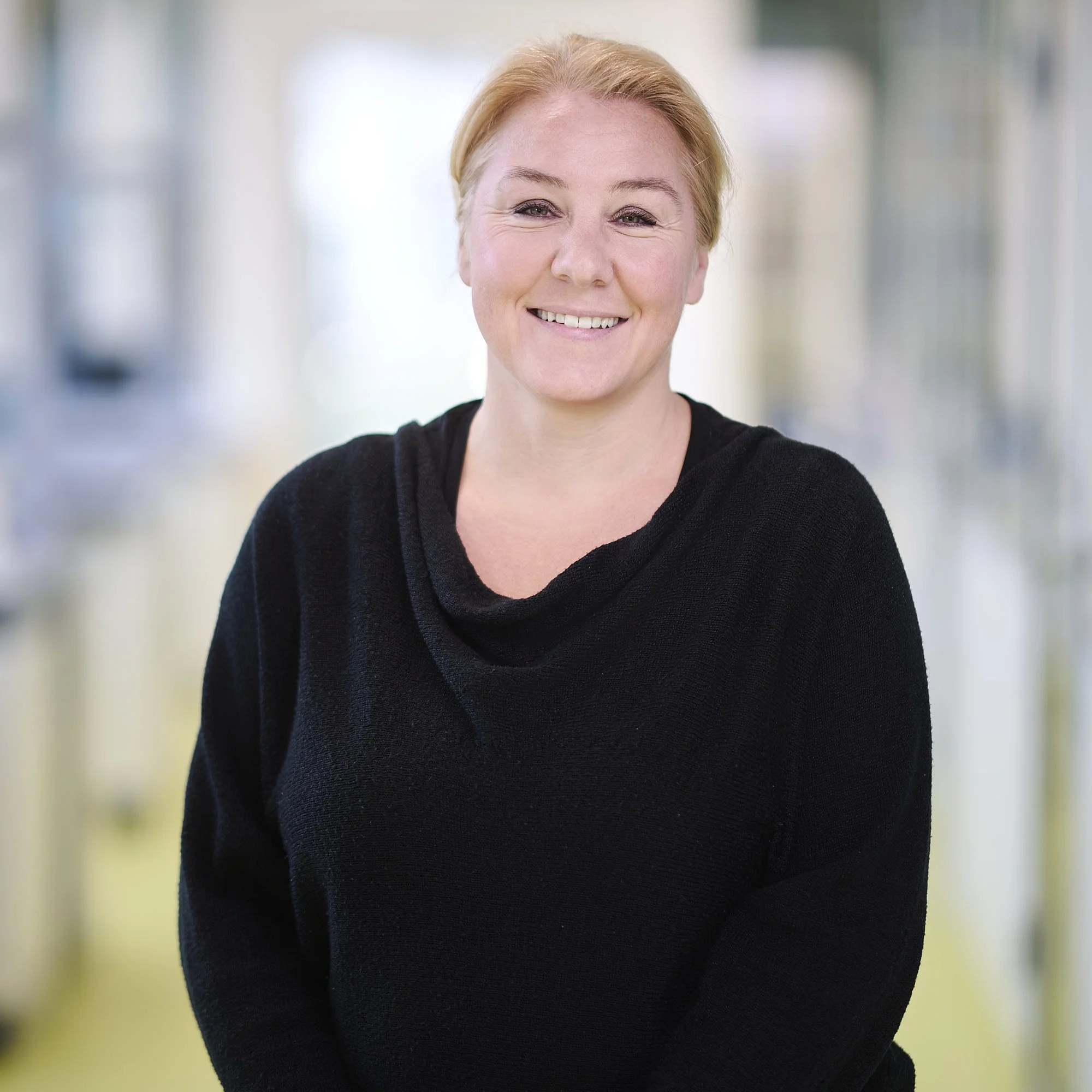 Smiling woman with short, red hair in a black sweater standing in a brightly lit modern indoor space.