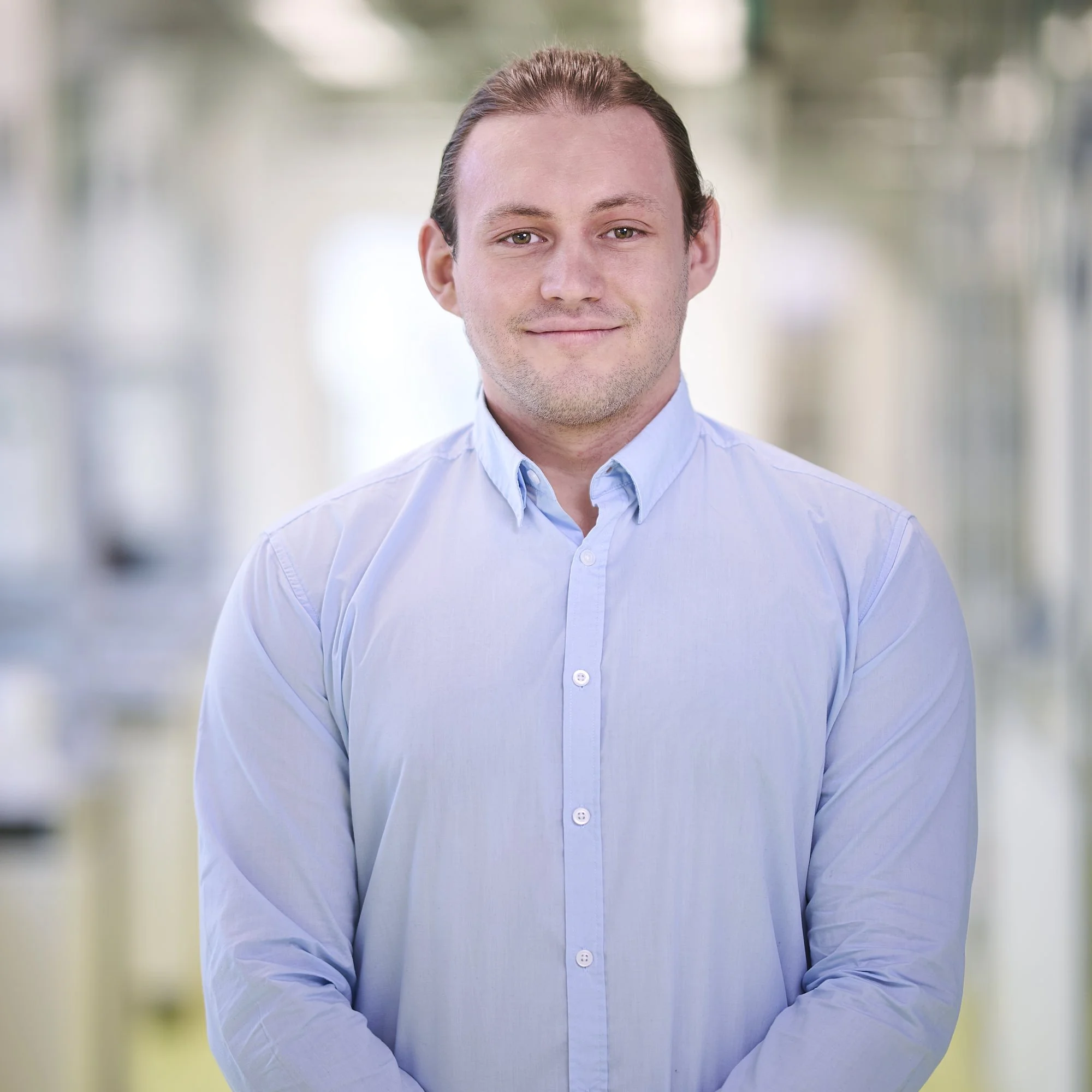 Portrait of a young man with brown hair and fair skin, wearing a light blue button-up shirt, standing in a softly blurred indoor setting with natural light.