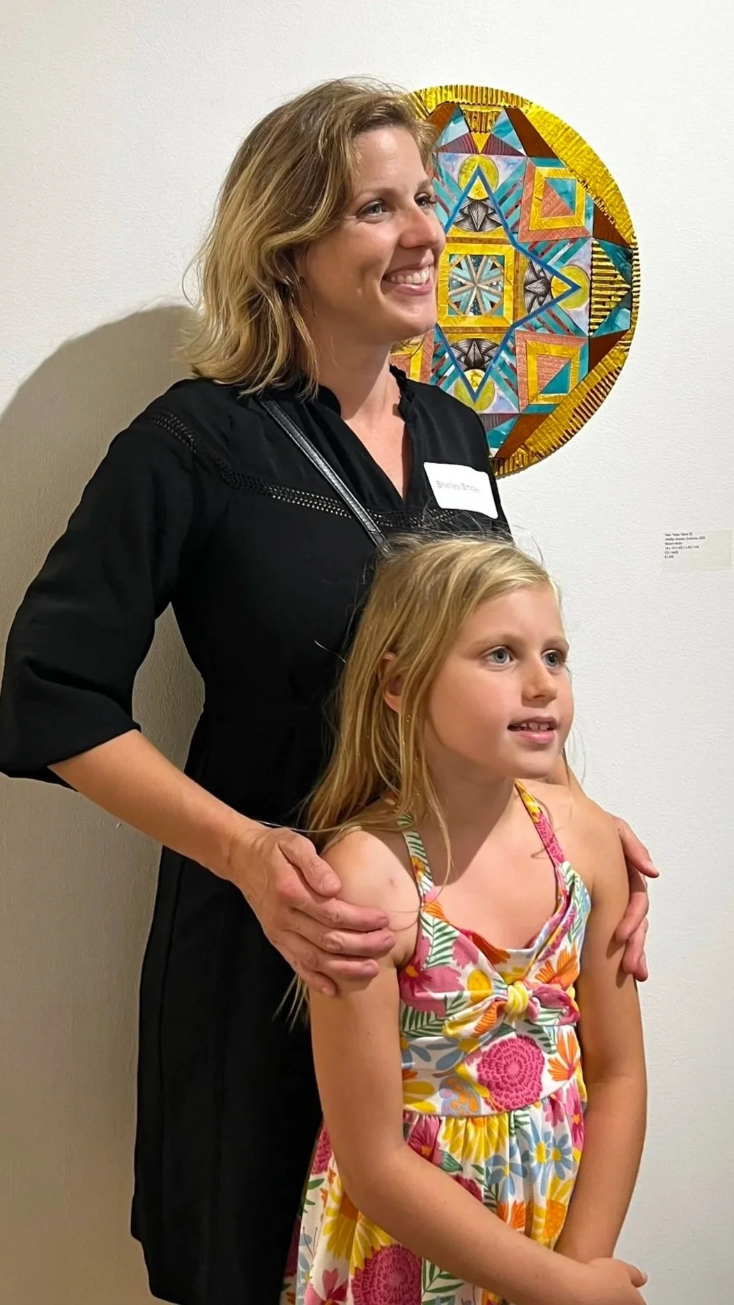 A woman and a young girl stand together in front of a white wall with colorful geometric artwork. The woman has shoulder-length blonde hair and is wearing a black dress, smiling. The girl with long blonde hair wears a colorful floral dress and looks 