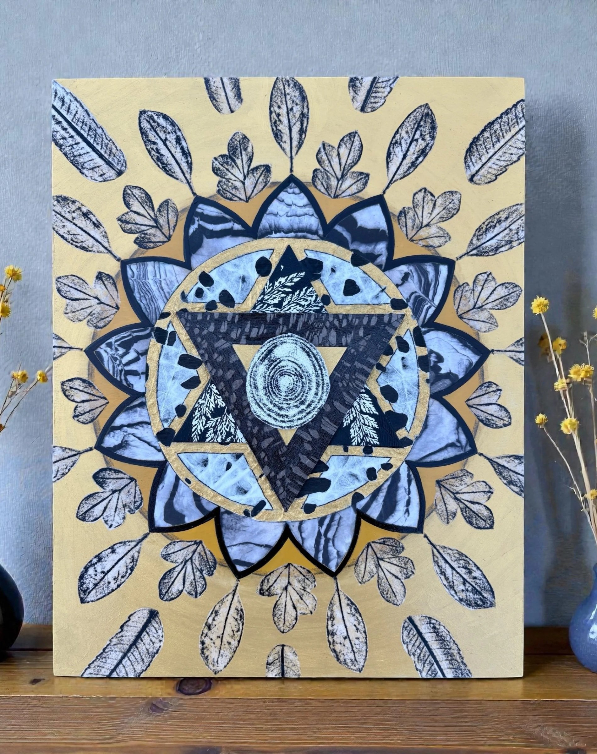 Decorative artwork featuring a geometric design with a central tree ring pattern, surrounded by a methane symbol and a circle, with black and white leaf motifs and a gold background. Flanked by two small vases with dried yellow flowers on a wooden su