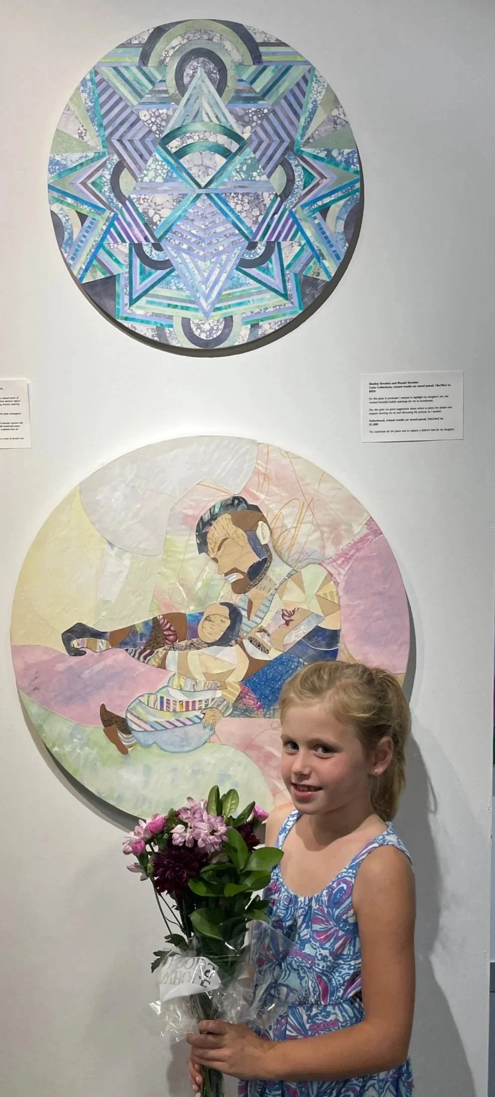 Young girl with blonde hair wearing a blue patterned dress holding a bouquet of flowers, standing in front of two circular art pieces, one depicting a mandala-style geometric design and the other a quilt-like piece with a figure, displayed in an art 