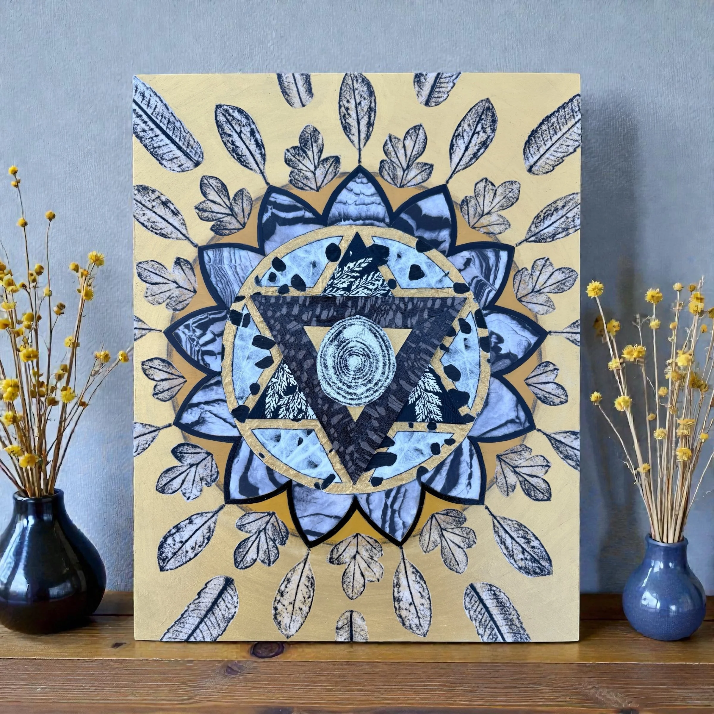 Decorative artwork featuring a geometric design with a central tree ring pattern, surrounded by a methane symbol and a circle, with black and white leaf motifs and a gold background. Flanked by two small vases with dried yellow flowers on a wooden su