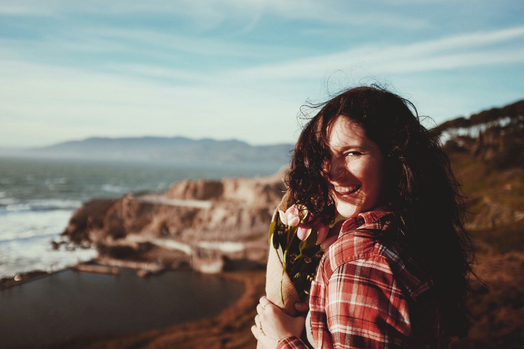 A woman with curly dark hair smiling and holding a bouquet of pink tulips, standing outdoors with a coastal landscape and cliffs in the background.
