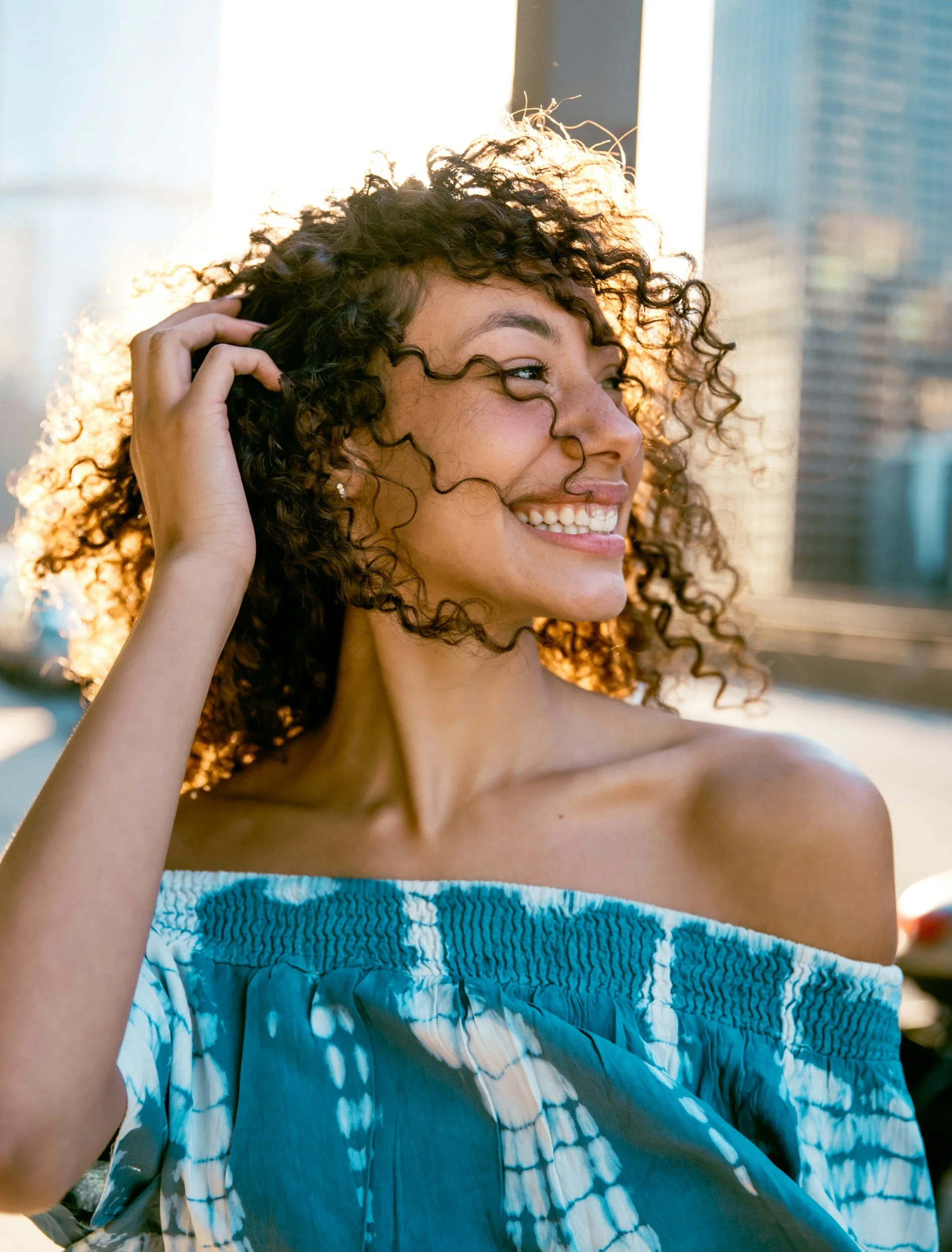 A woman with curly hair smiling and touching her hair, wearing an off-the-shoulder blue patterned top, with city buildings and sunlight in the background.
