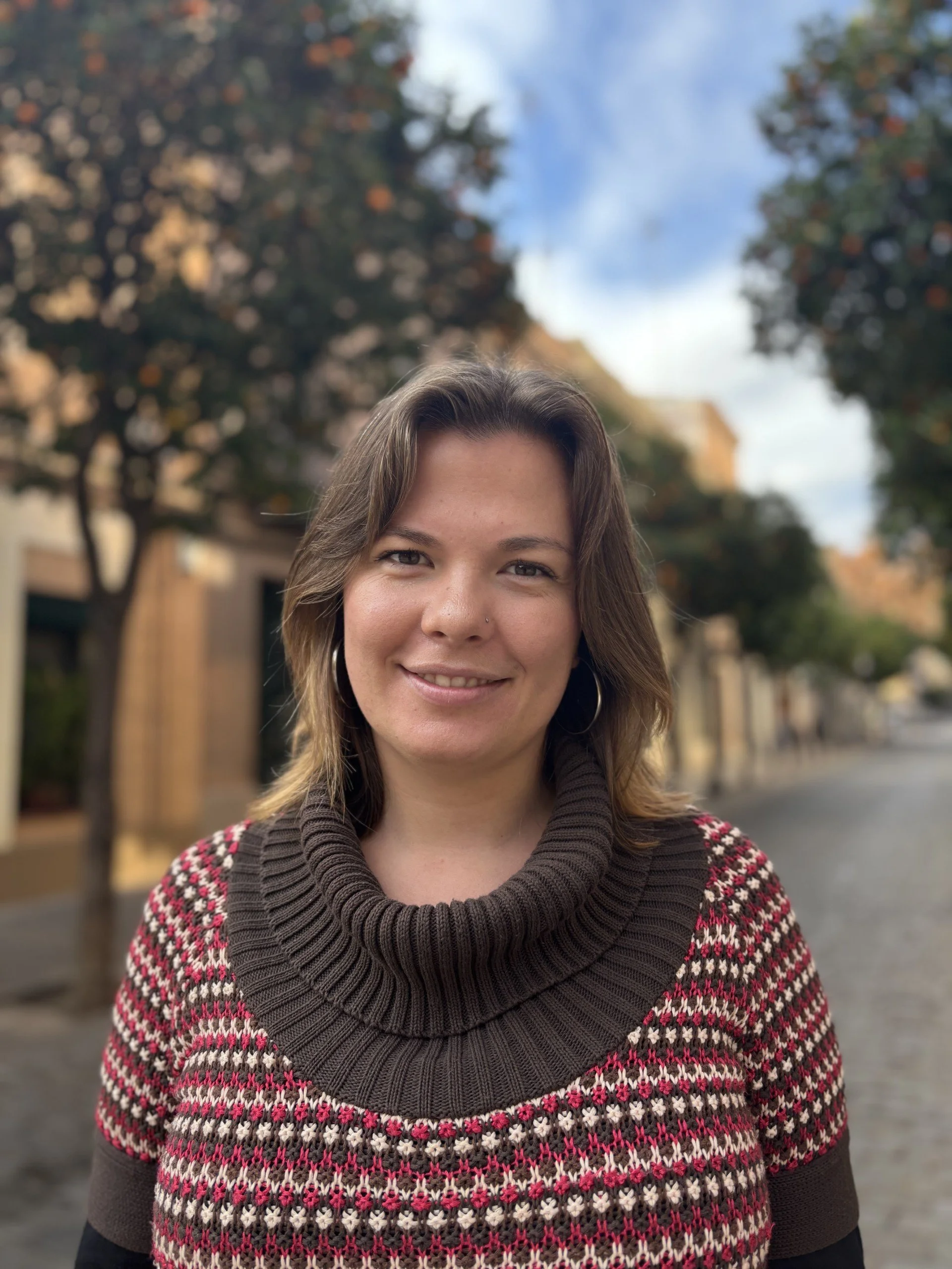 A woman with shoulder-length brown hair, wearing a patterned sweater, smiling outdoors on a street lined with trees and buildings, during a partly cloudy day.