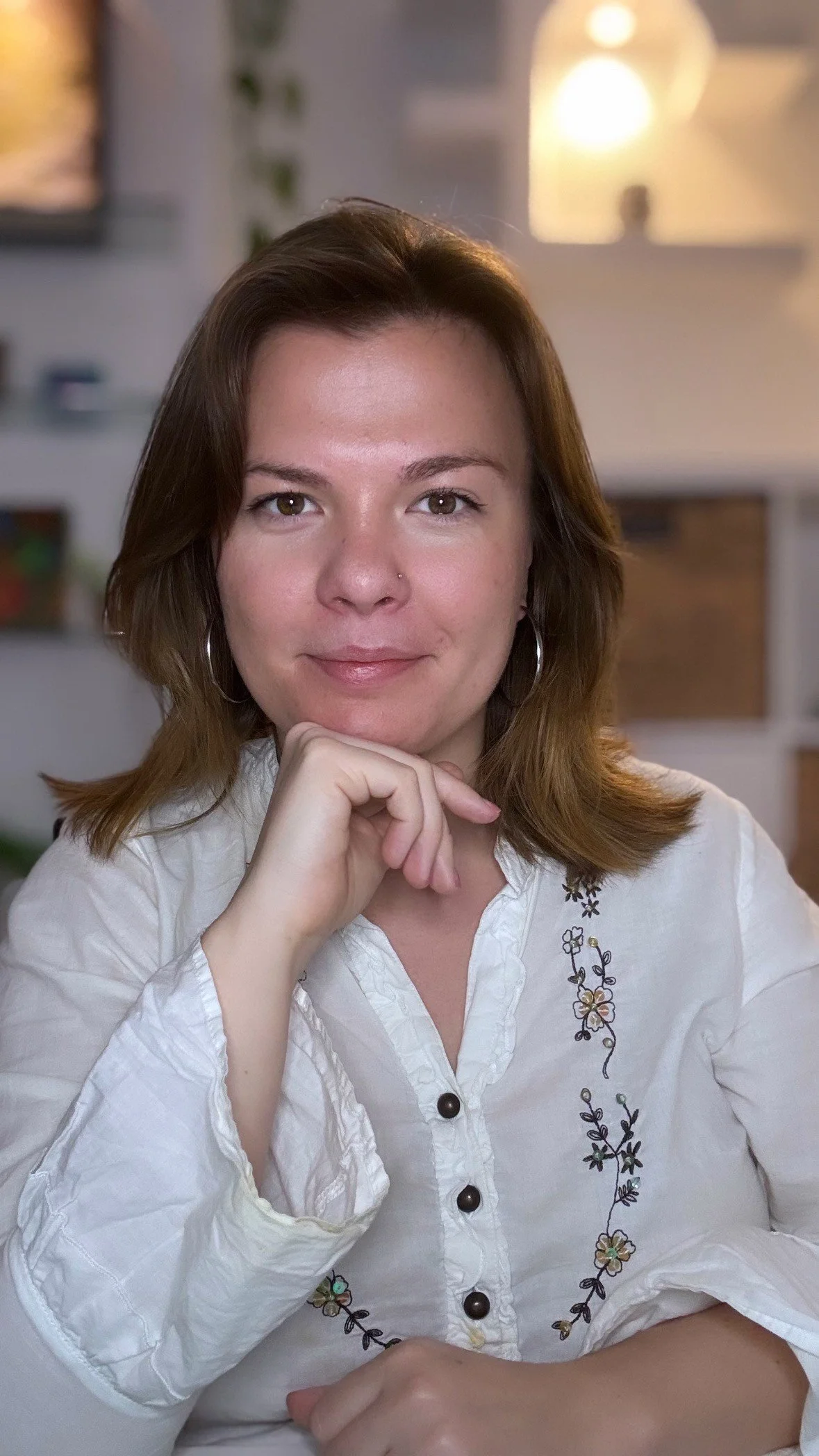 A woman with shoulder-length brown hair, wearing hoop earrings and a white embroidered blouse, sits at a table indoors with her chin resting on her hand, smiling softly at the camera.