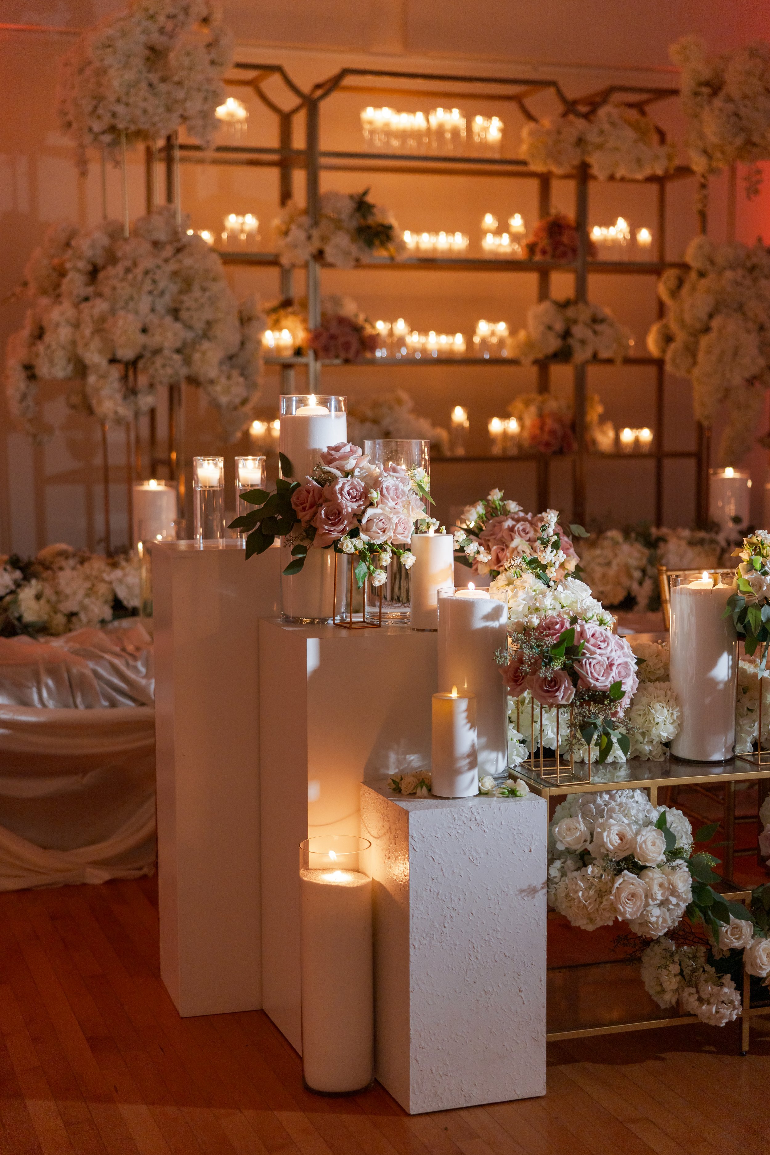 Elegant wedding decor featuring candles and floral arrangements with pink and white roses, set against a backdrop of shelves with candles and large flower clusters.