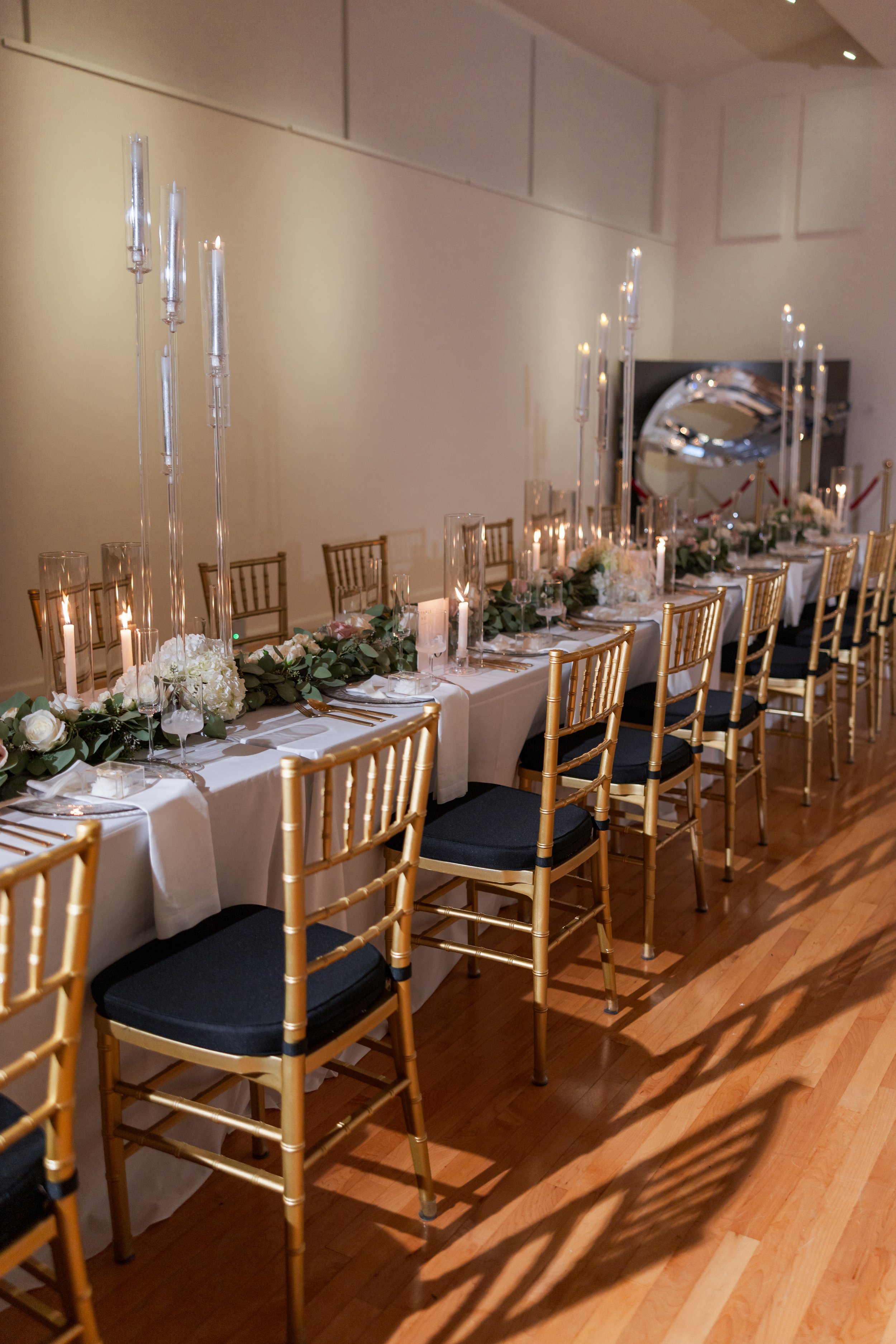 Elegant banquet table decorated with white flowers, green foliage, and tall glass candle holders with lit candles, set with gold chairs with black cushions.