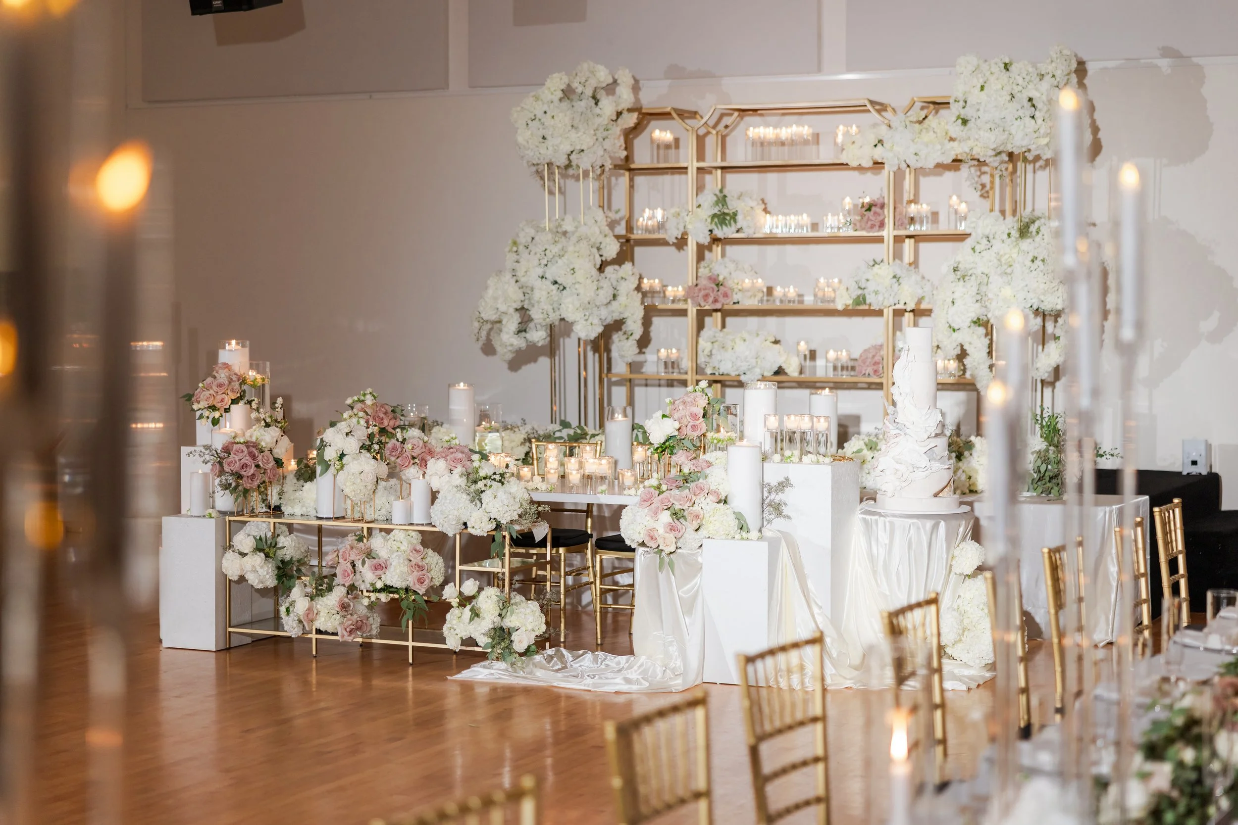 Elegant wedding reception decor with white and blush pink flowers, candles, and gold accents on a wooden floor, with a floral backdrop.
