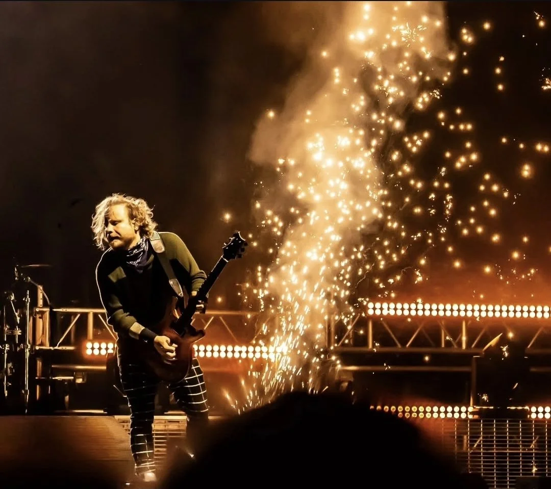 A musician with long, blonde, curly hair playing an electric guitar on stage amid sparks and bright lights during a concert.