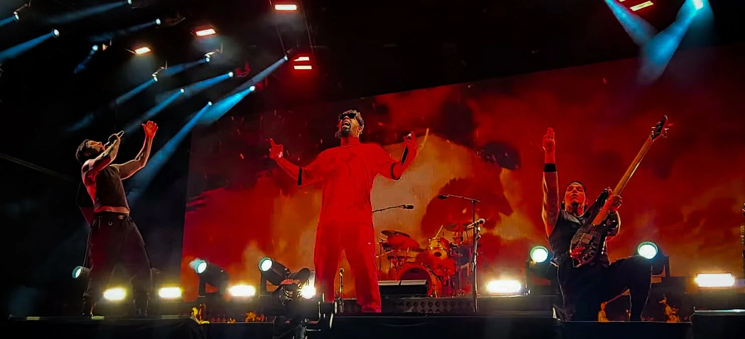 A band performing on stage with a background of fire and orange flames, featuring a vocalist, a guitarist, and a drummer.