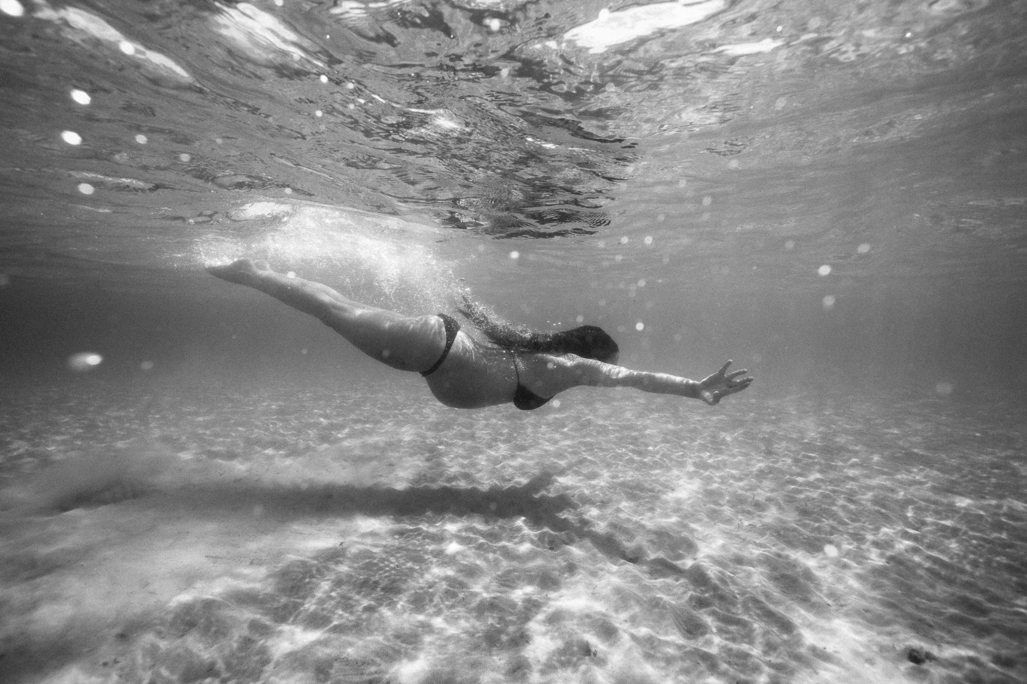 A woman swimming underwater in a bikini, reaching out with one arm, in clear water with the sandy bottom visible.
