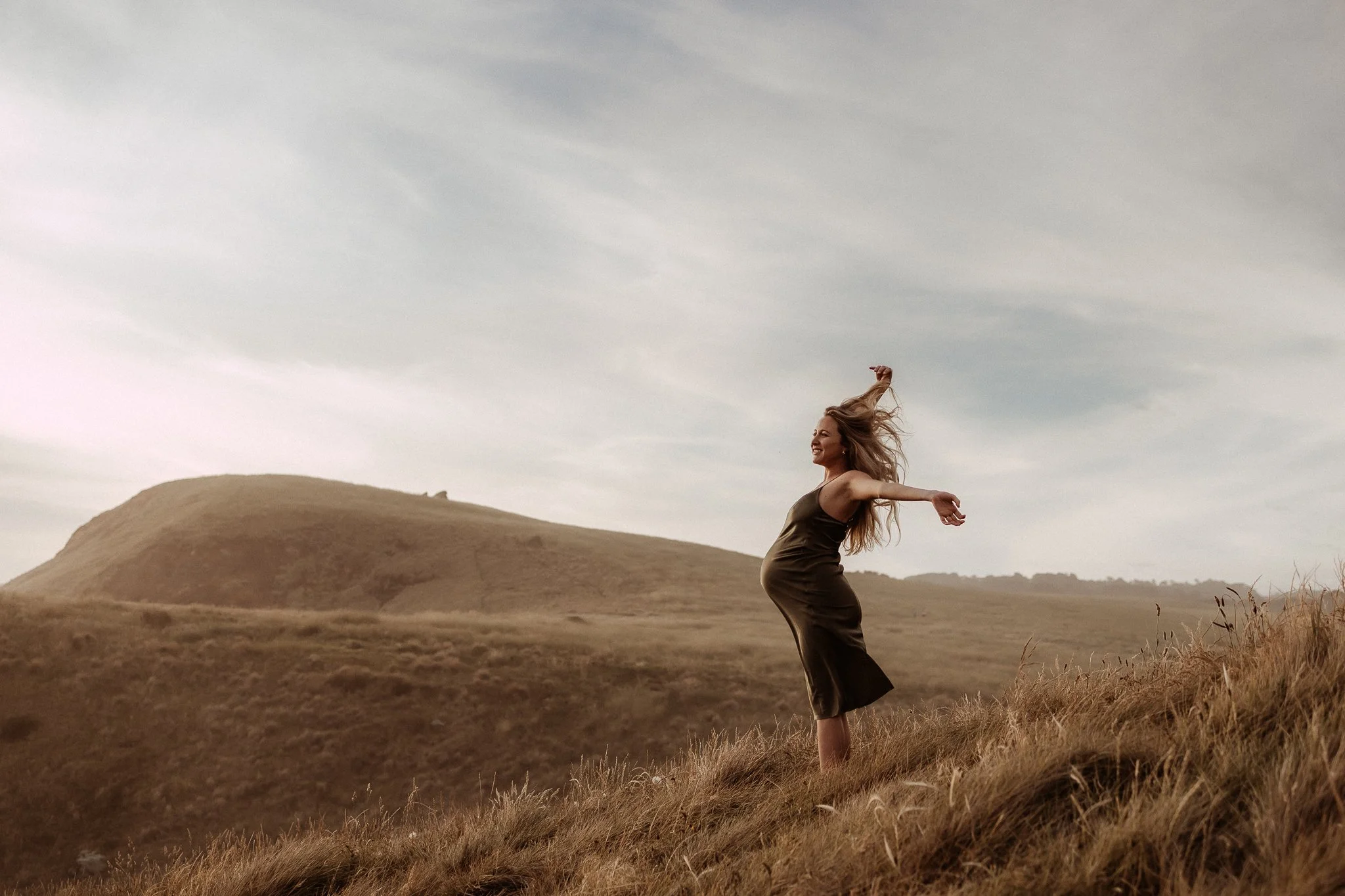A woman in a dark dress standing on a grassy hillside with arms outstretched and hair blowing in the wind, with hills and a cloudy sky in the background.