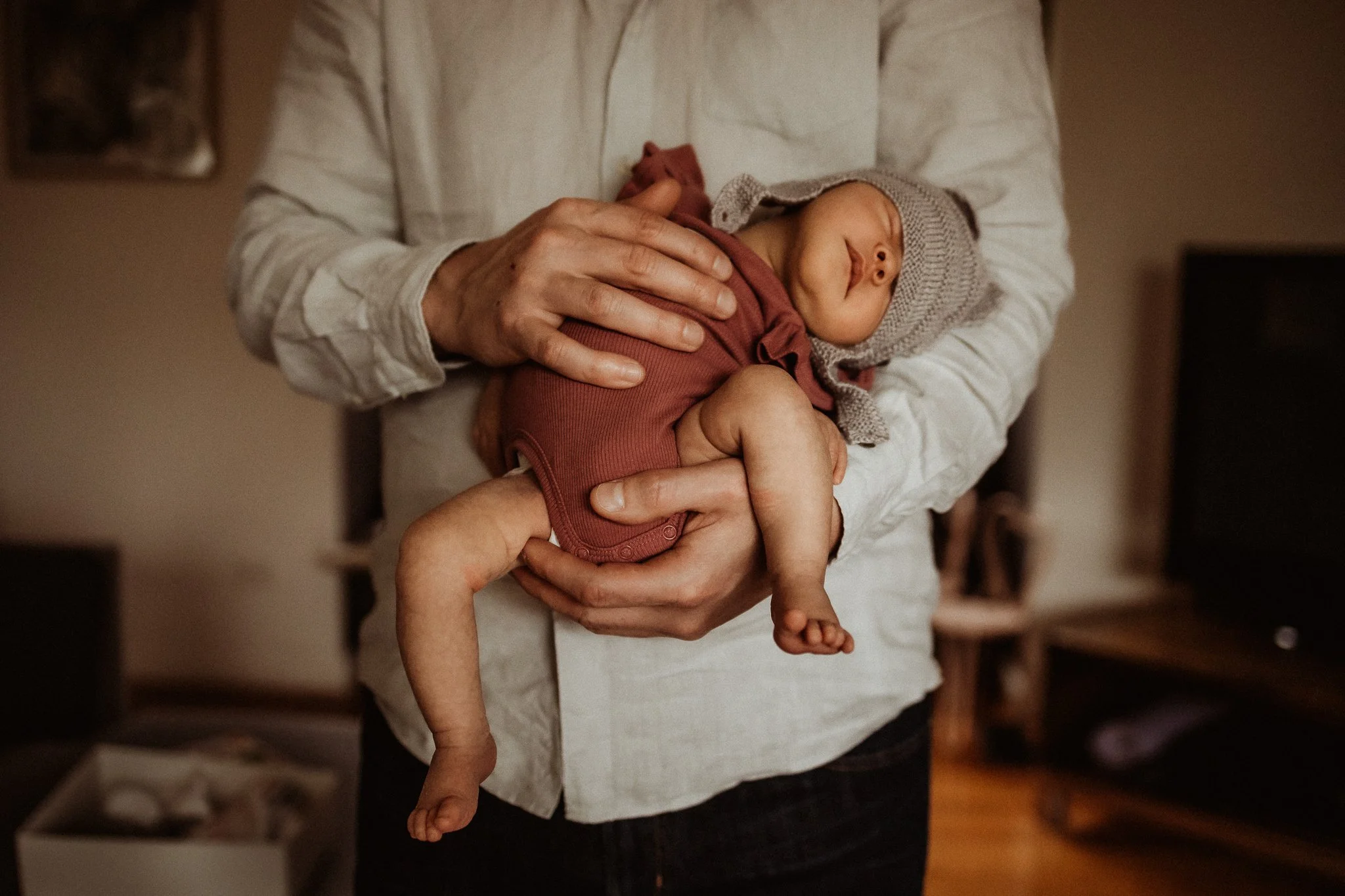 Person holding a sleeping baby dressed in a brown outfit and gray hat indoors.