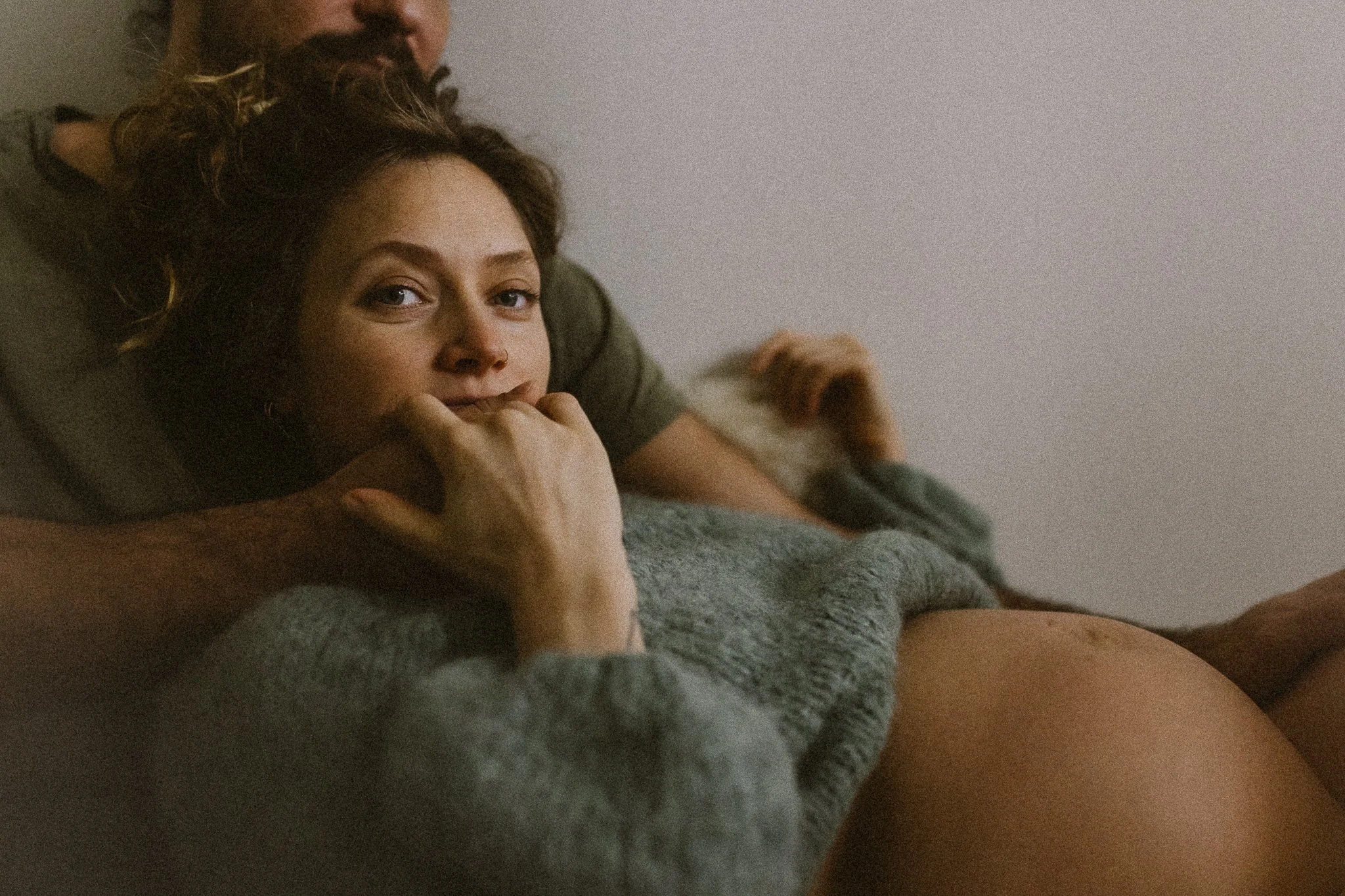 A woman lying down with her head on a man's shoulder, looking at the camera, with her hand near her mouth. They are in a relaxed setting with a plain background.