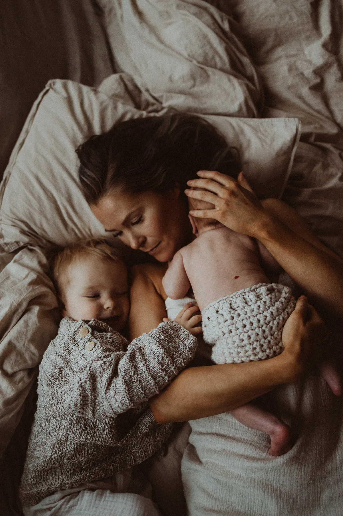 A woman with long dark hair, a young boy, and a baby lying together on a bed. The woman is cuddling the baby, and they all appear to be sleeping peacefully.