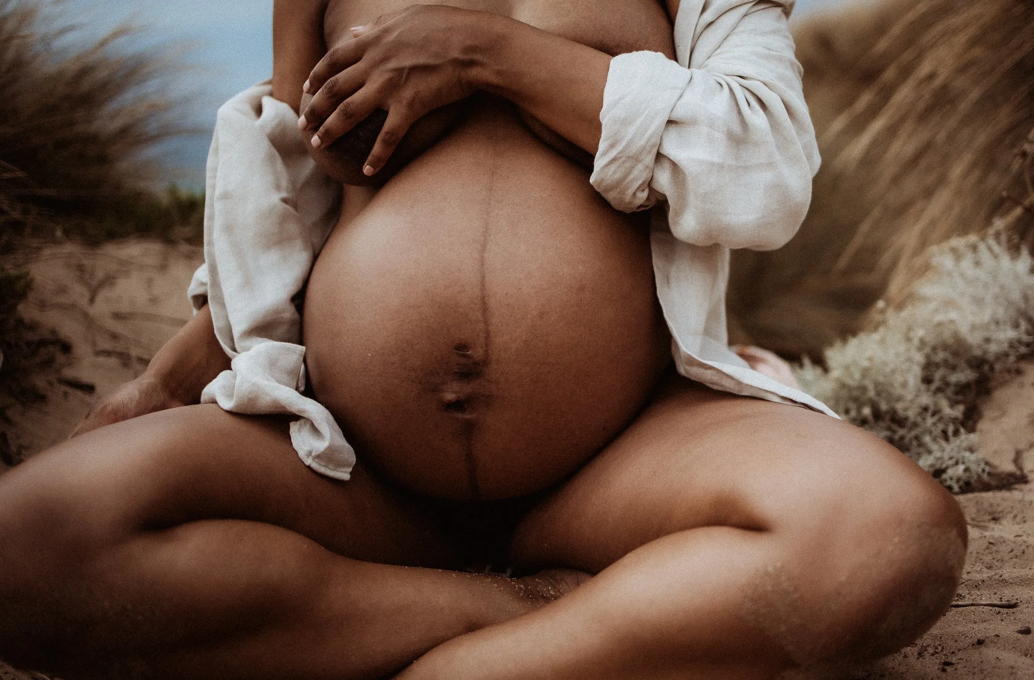 A close-up of a pregnant woman sitting on the sand at the beach, wearing a white shirt and holding her belly.