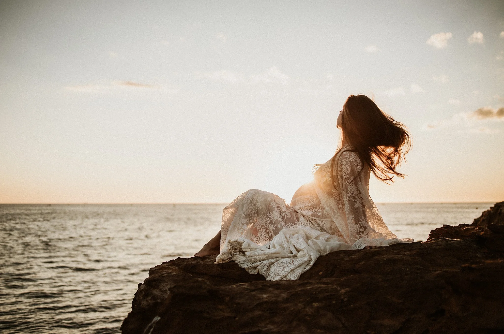 A woman in a lace dress sitting on a rocky shore during sunset, with her hair flowing and her face turned away from the camera.