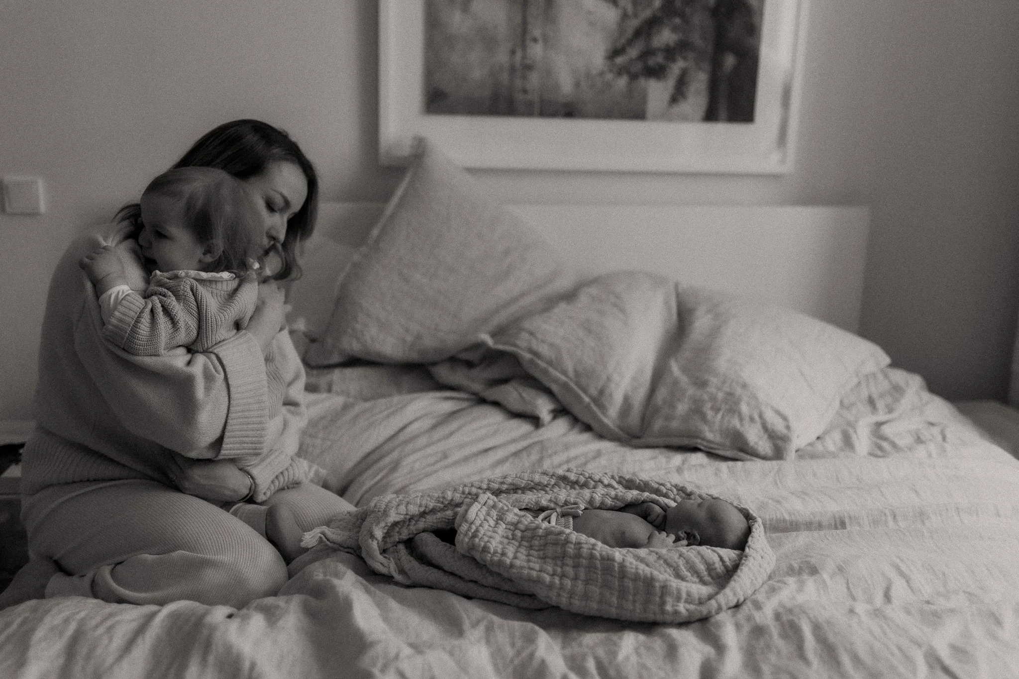 A woman sitting on a bed holding a baby in her arms, with another infant lying in a blanket on the bed.