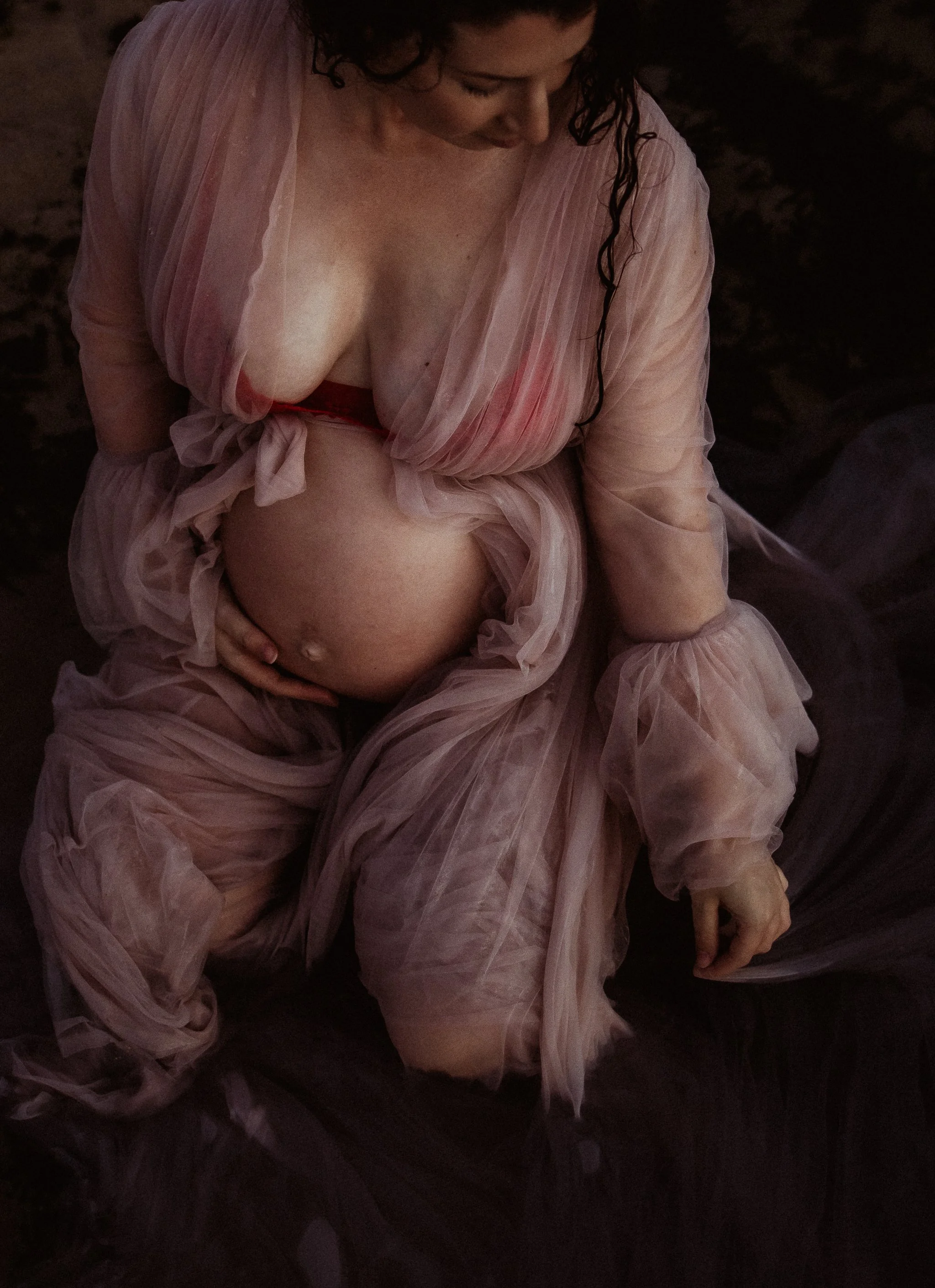A pregnant woman sitting down, wearing a pink sheer cover-up and red lingerie, with her hand resting on her belly and head bowed, in a dimly lit setting.