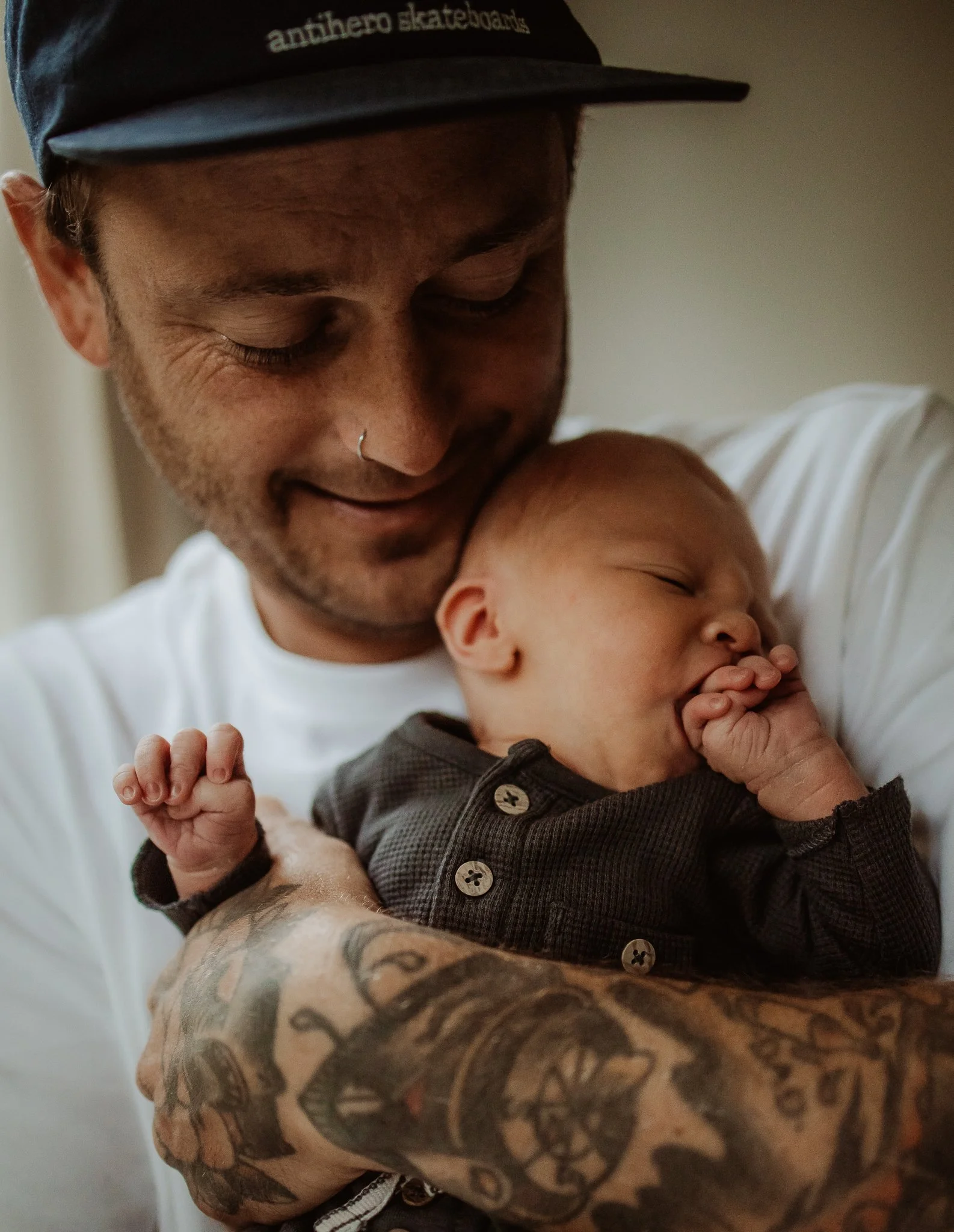 A man with tattoos on his arm and a nose piercing, wearing a baseball cap, holding a sleeping baby close to his chest.