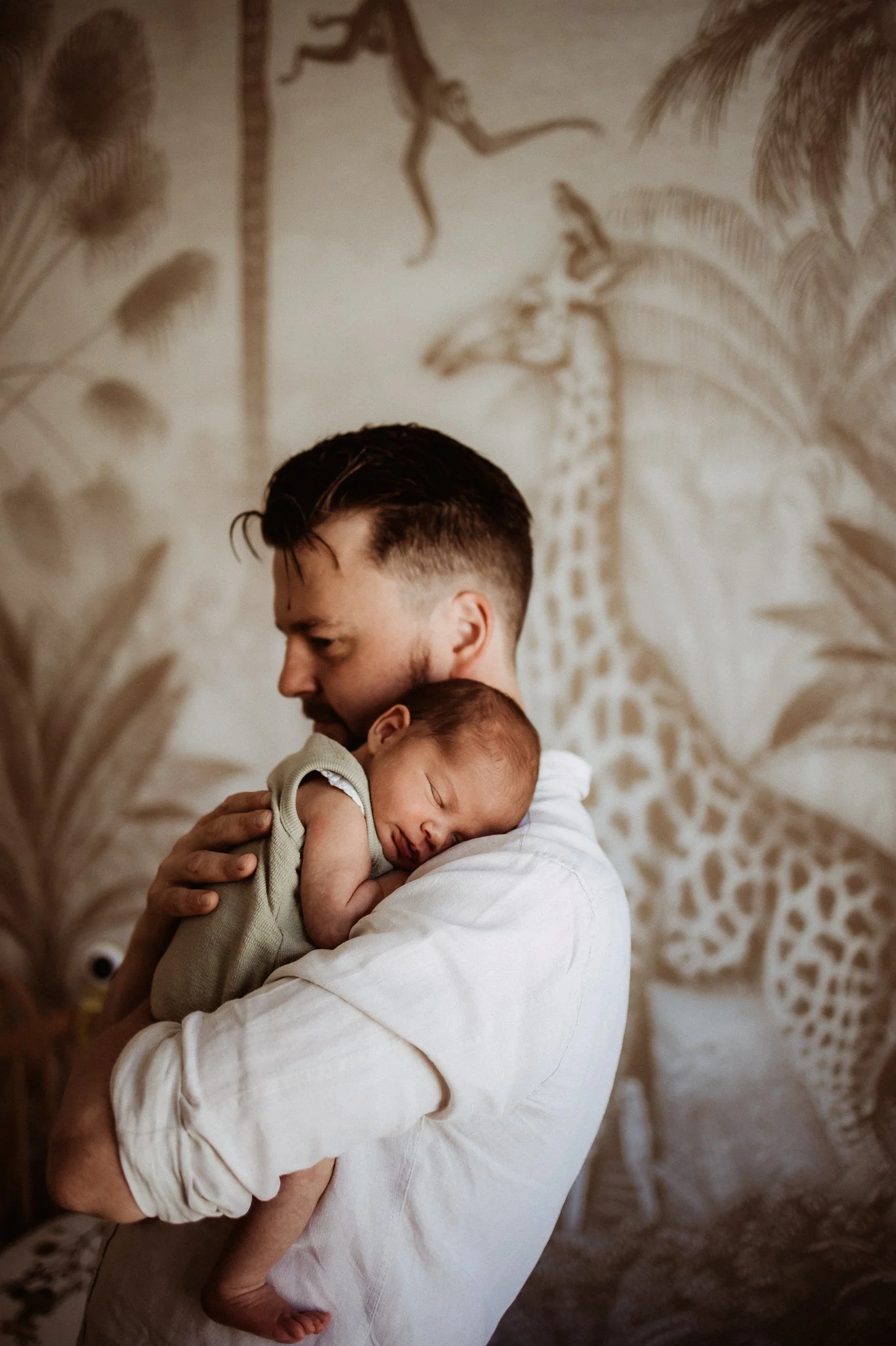 A man holding a sleeping baby in front of a zebra and giraffe mural.