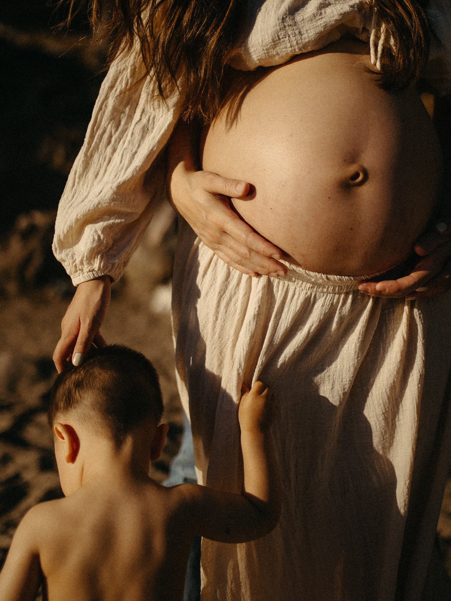 Pregnant woman in a light dress holding her belly with a young child standing next to her.
