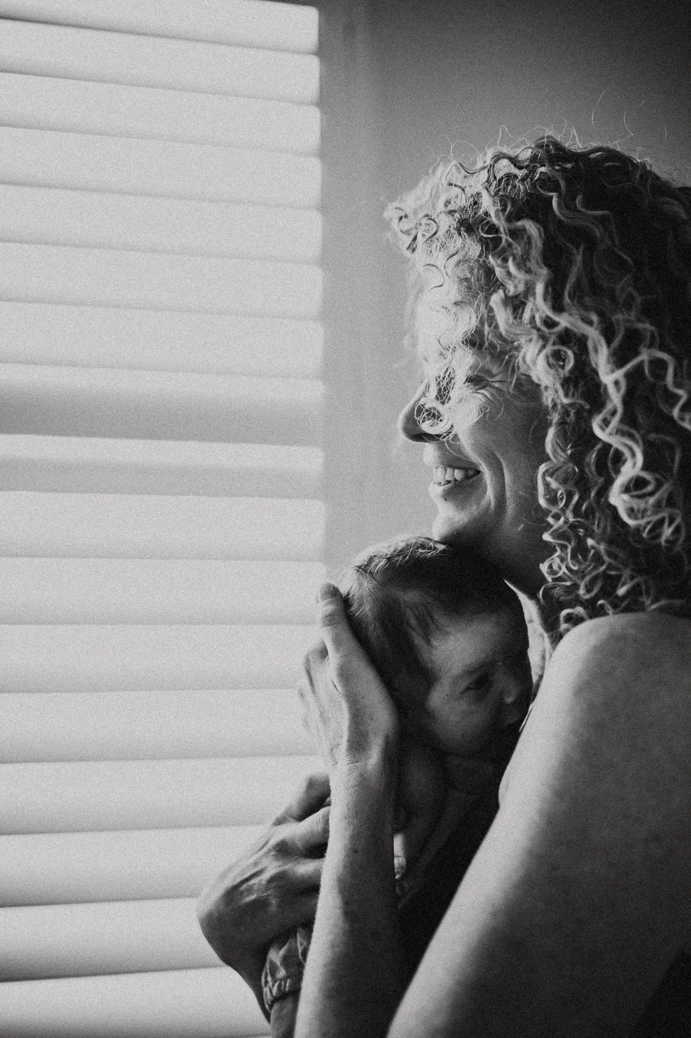 A black and white photo of two people hugging, one with curly hair and the other with long straight hair, near a window with blinds.