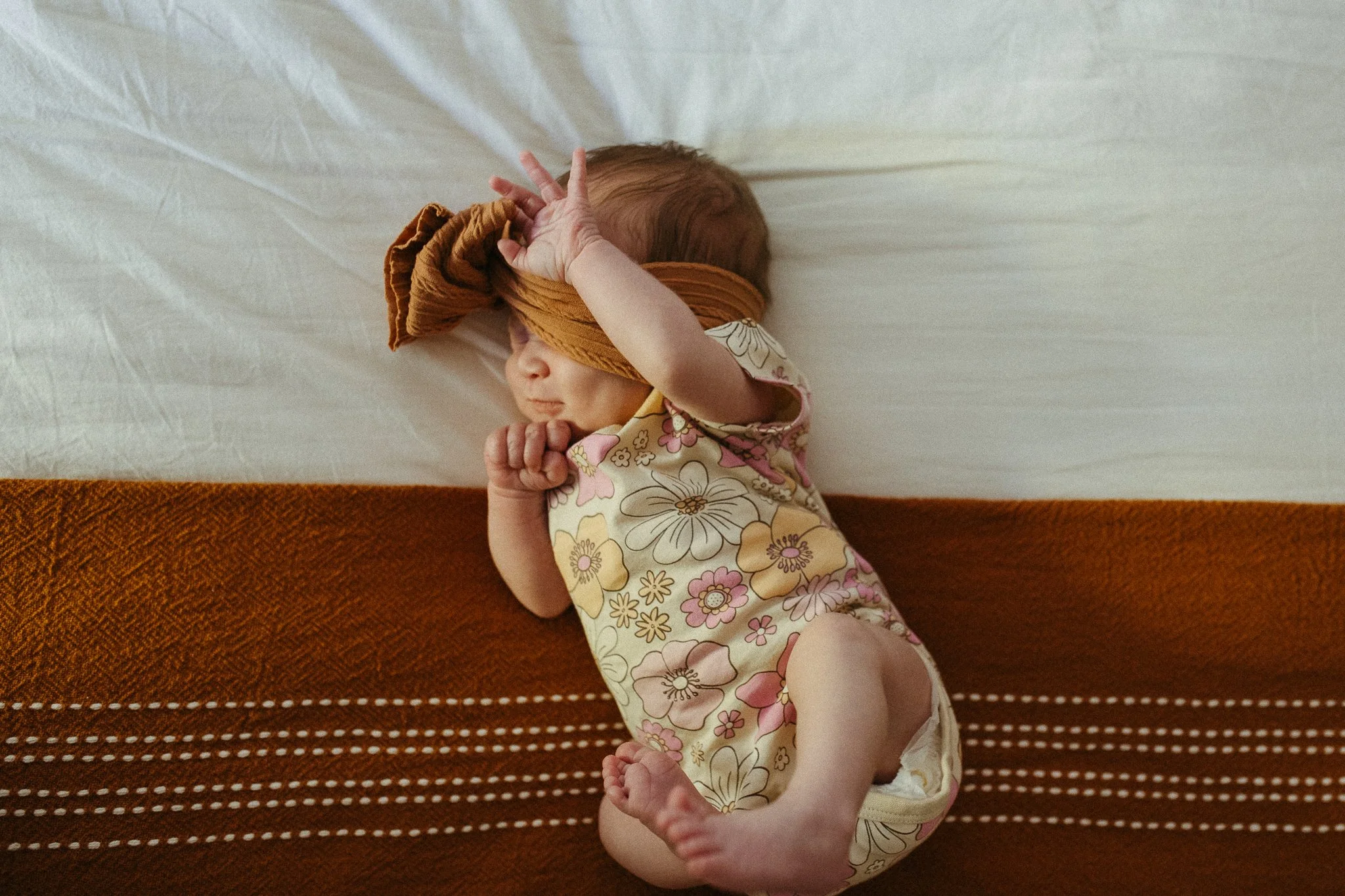 A baby girl sleeping on a bed, wearing a floral dress and a brown headscarf, with a brown blanket partially covering her.