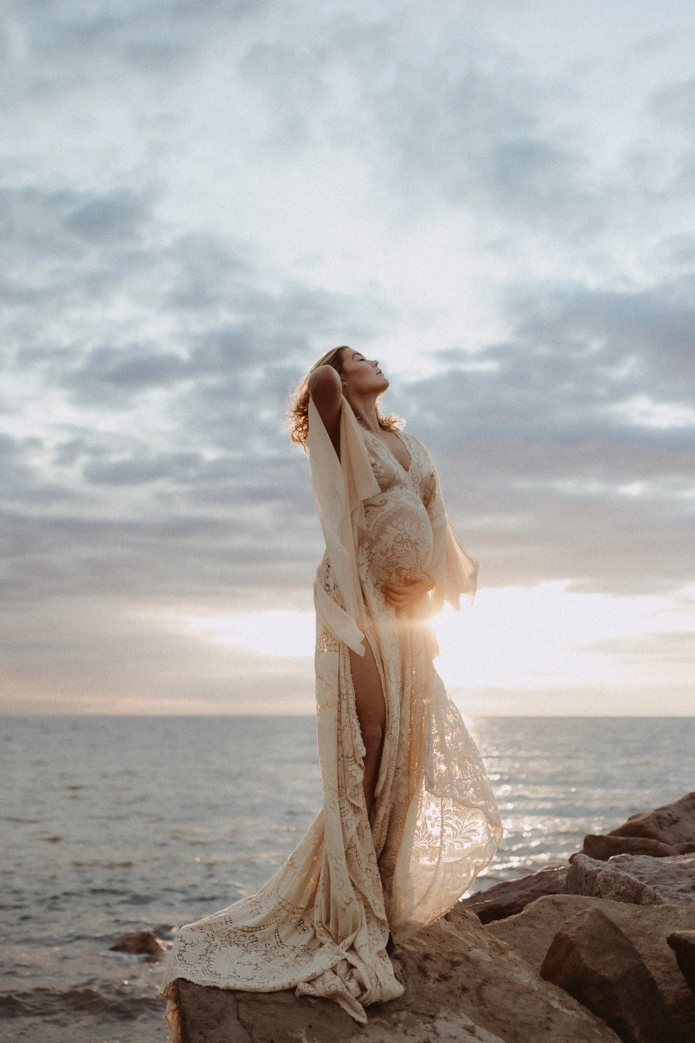 A pregnant woman in a flowing lace dress standing on rocks by the ocean during sunset, with her hand on her belly and her eyes closed, enjoying the moment.