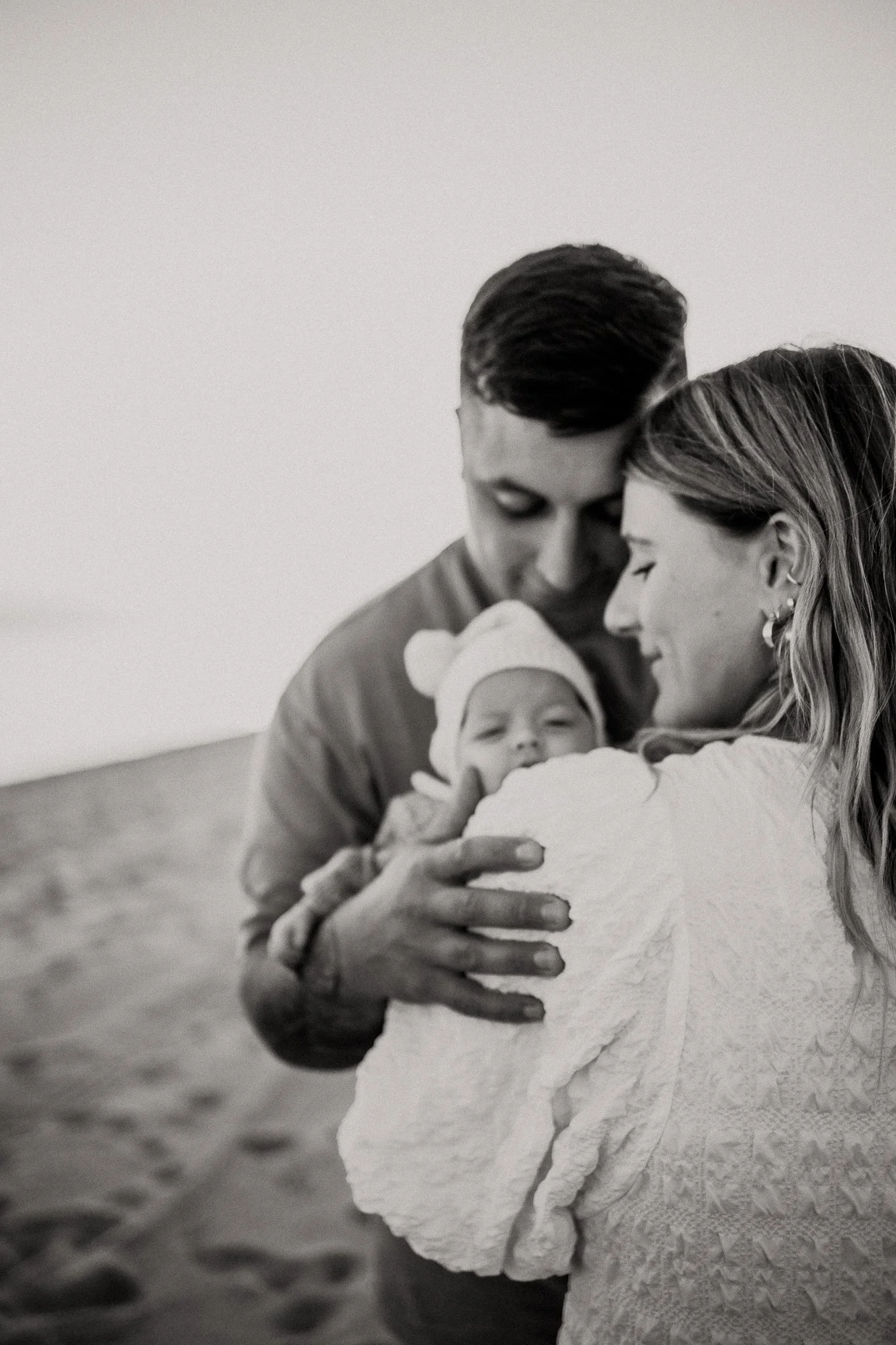 A woman with long hair embraced by a man with short hair, holding a baby wrapped in a blanket, in a black and white photo.