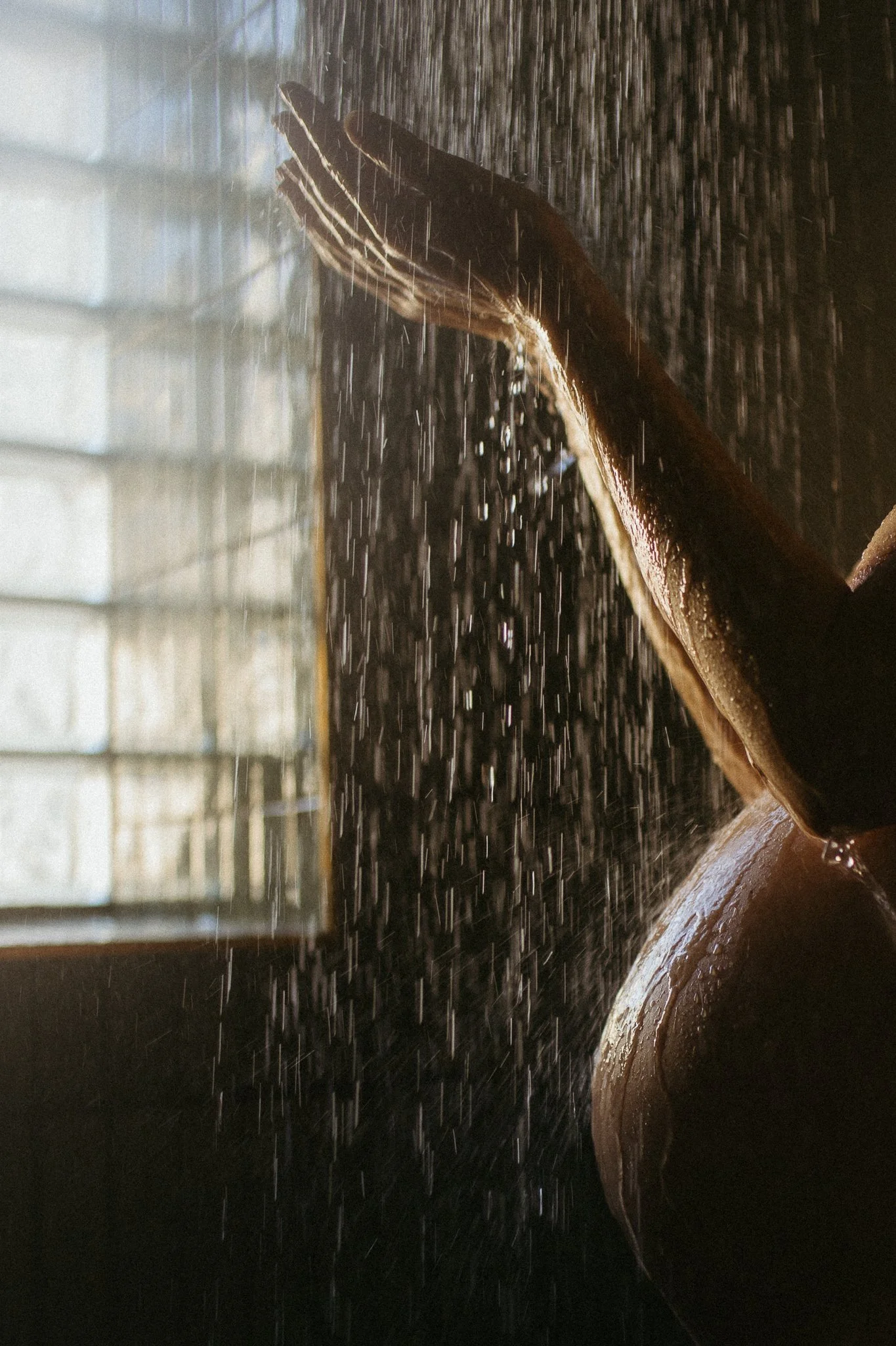 A pregnant mother's hand and arm raised under falling water in a dimly lit shower.