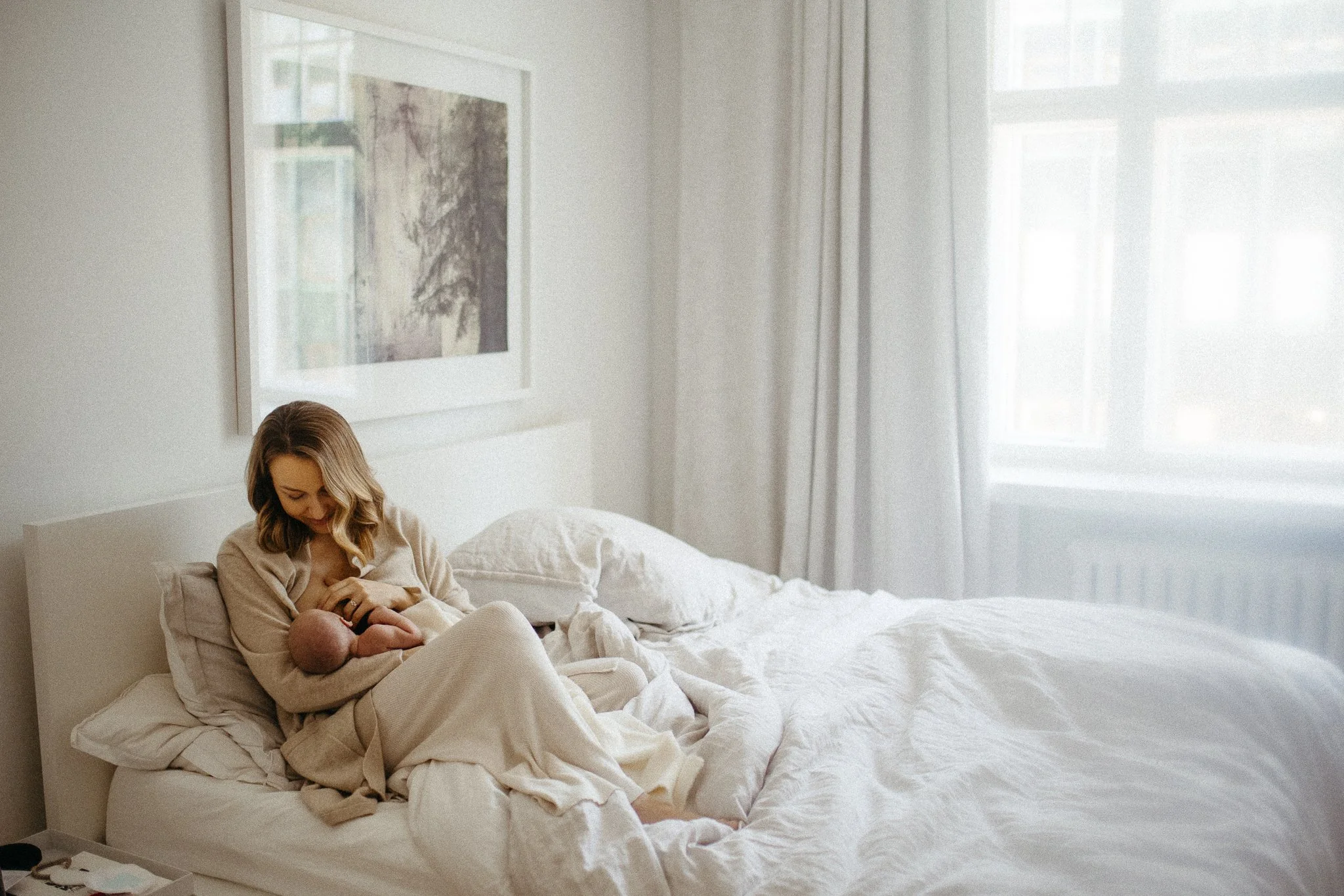 A woman in a beige robe sitting on a bed, breastfeeding a newborn baby in a bright, minimalistic bedroom with white bedding and large windows.
