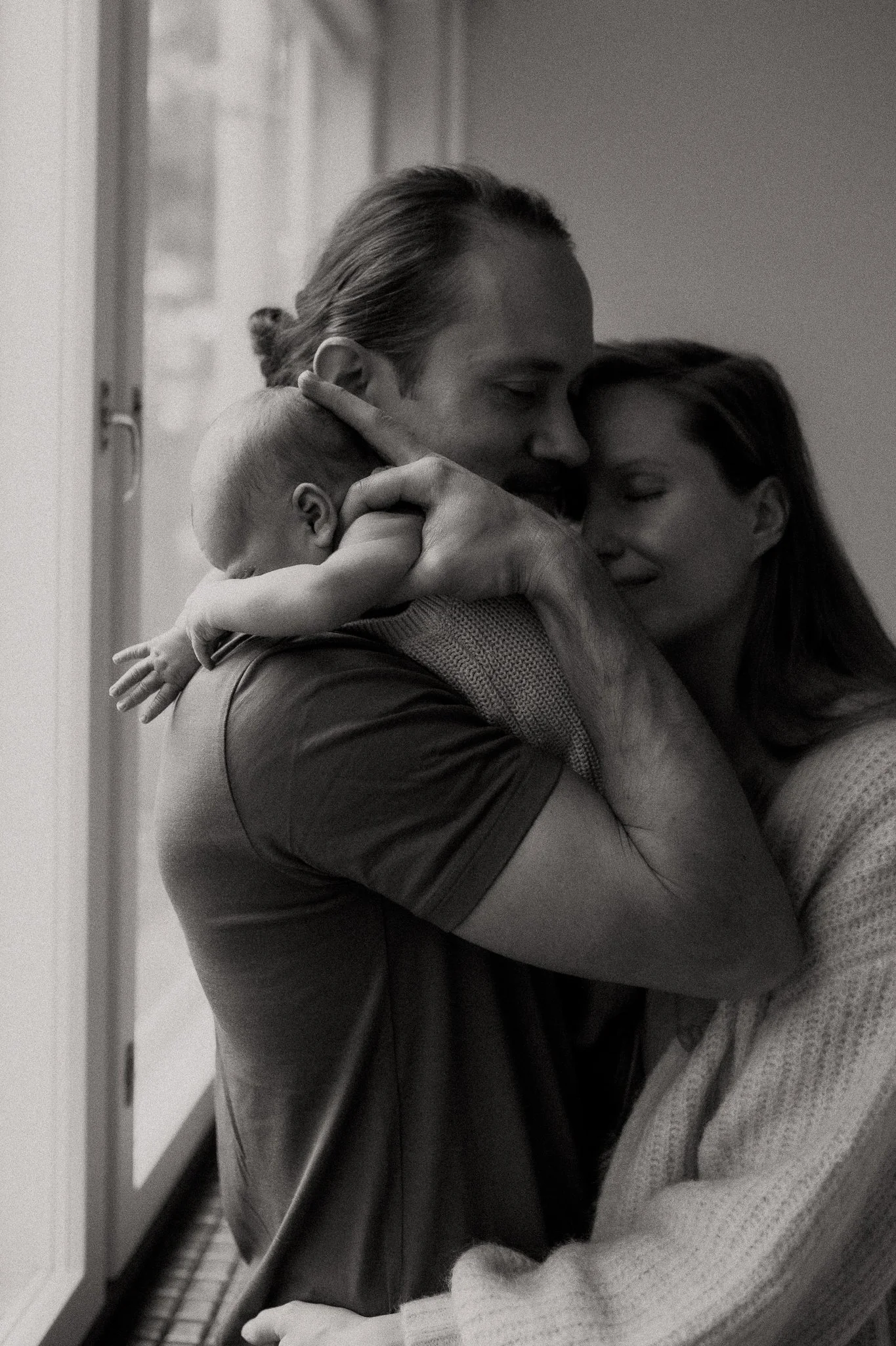 A black-and-white photo of a family embracing each other warmly. A man with long hair tied back is holding a woman and a baby, all with their eyes closed and smiling gently.