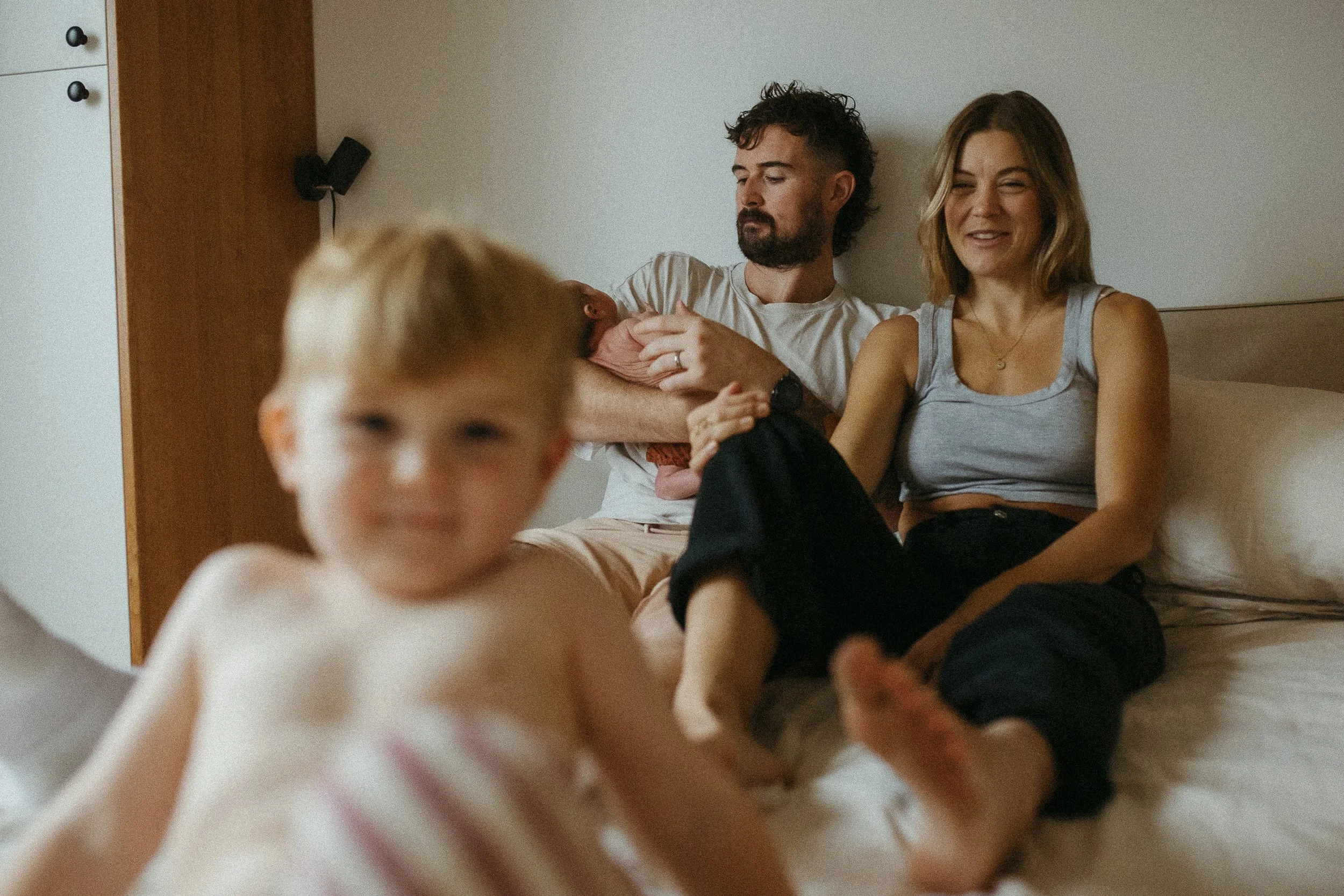 A family sitting on a bed, with a woman holding a baby, a man sitting beside her, and a young boy in the foreground. The woman is smiling, and the man appears content.