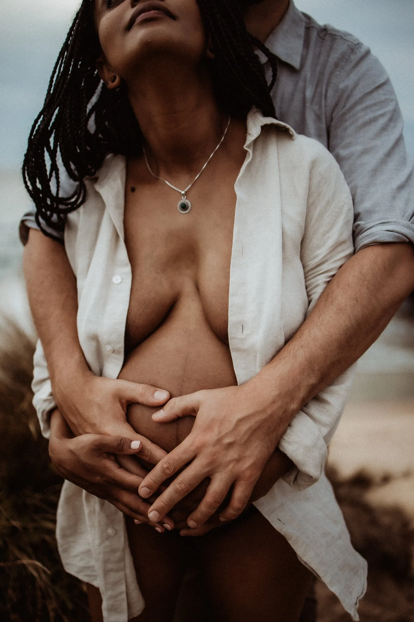 A close-up of a couple embracing on a beach, with the woman wearing an open shirt and a necklace, and both hands resting on her pregnant belly.