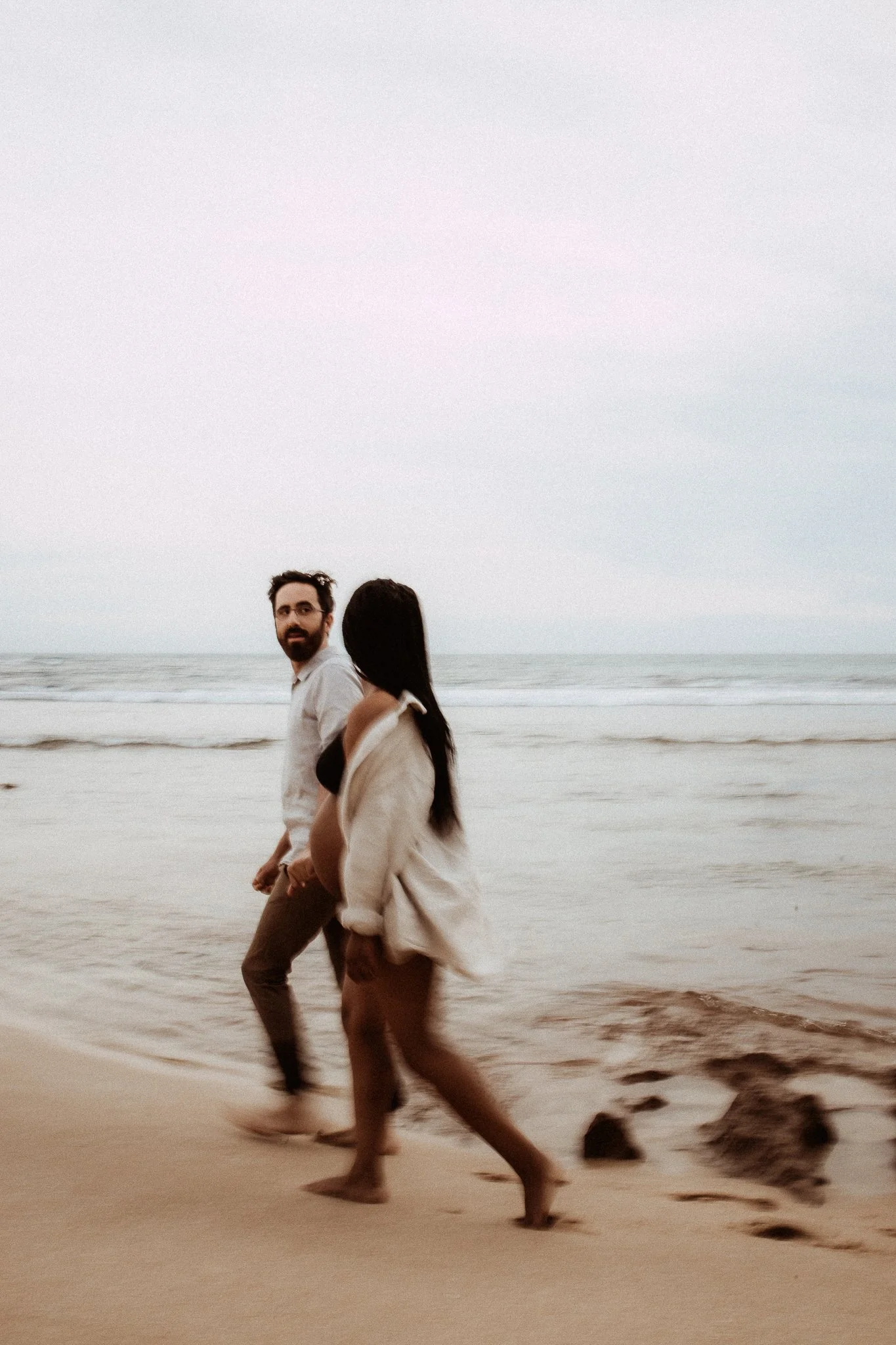 A man and woman walking barefoot on the beach near the water at sunset or sunrise, with the ocean in the background.