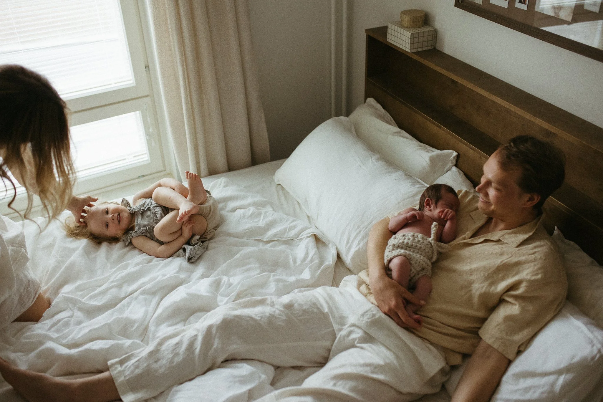 A family with two young children and two adults relaxing on a bed in a bedroom, softly lit by natural light through a window.