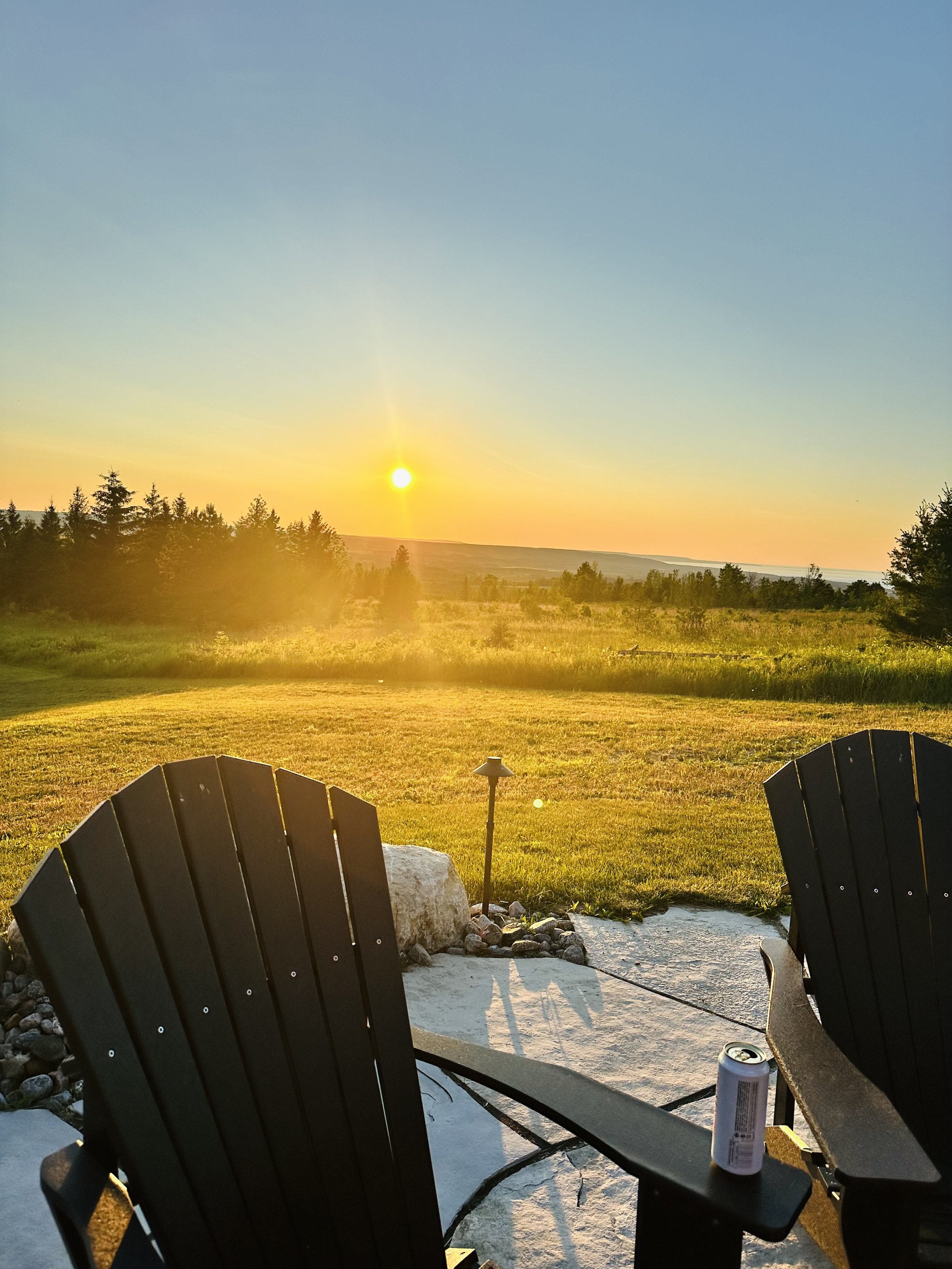 Two Adirondack chairs on a patio overlooking a grassy field with trees in the distance during sunset. A can is placed on one of the chair arms.