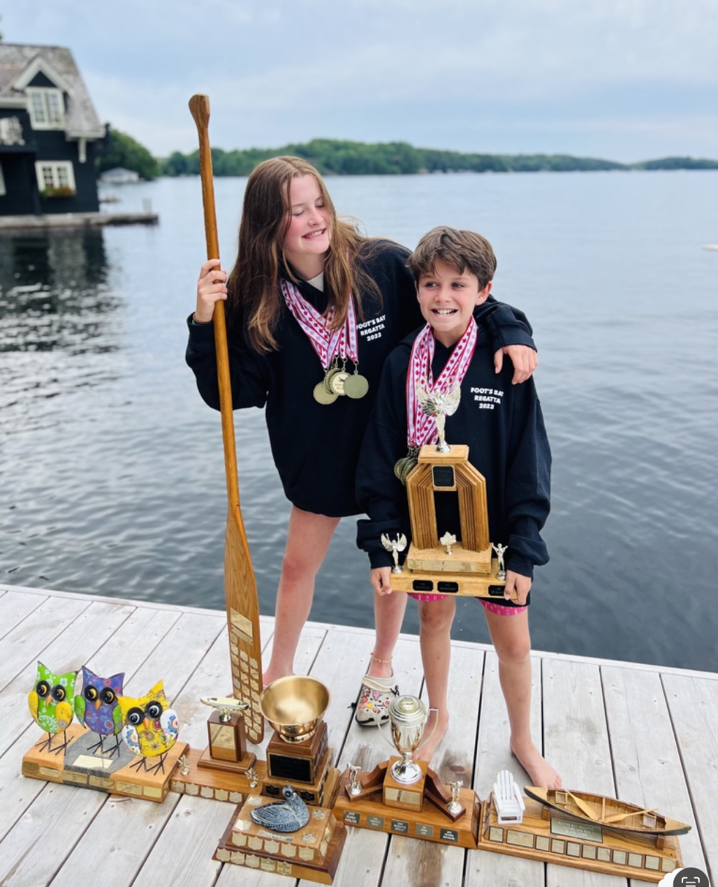 Two children on a dock wearing black hoodies adorned with medals, standing next to several wooden trophies with small owl decorations and plaques. They are beside a lake, with a house visible in the background.