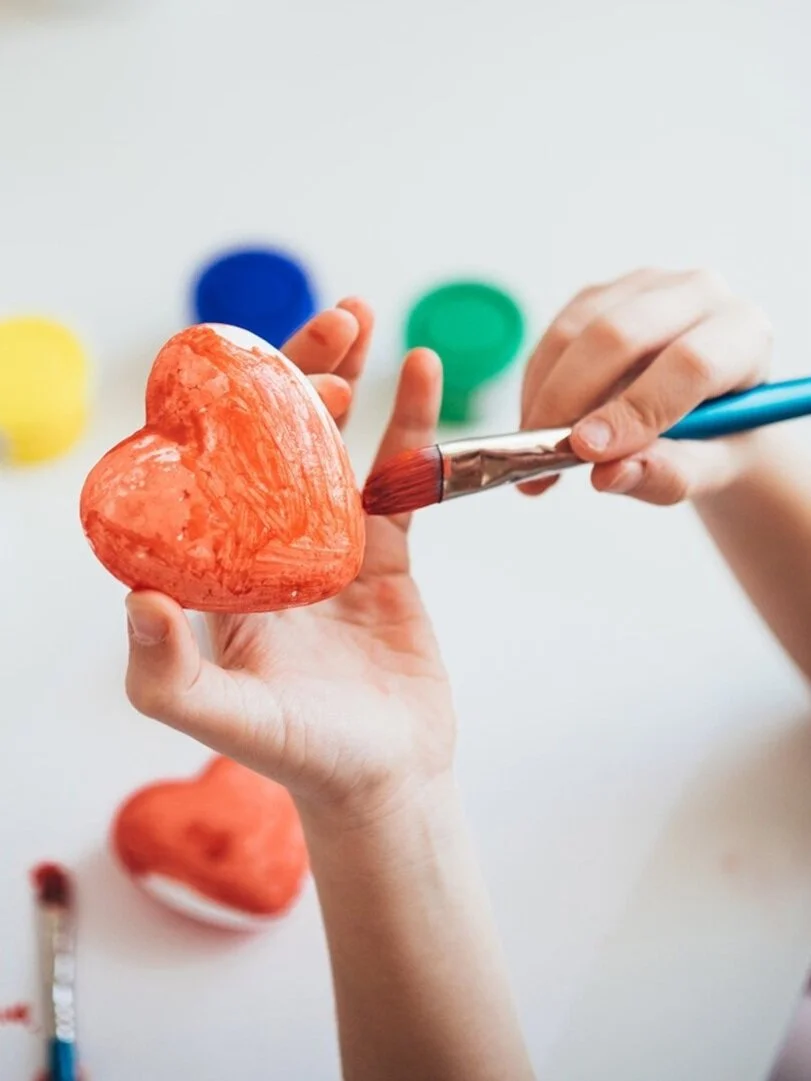 Person painting a red heart-shaped object with a paintbrush, with colorful paint jars in the background.