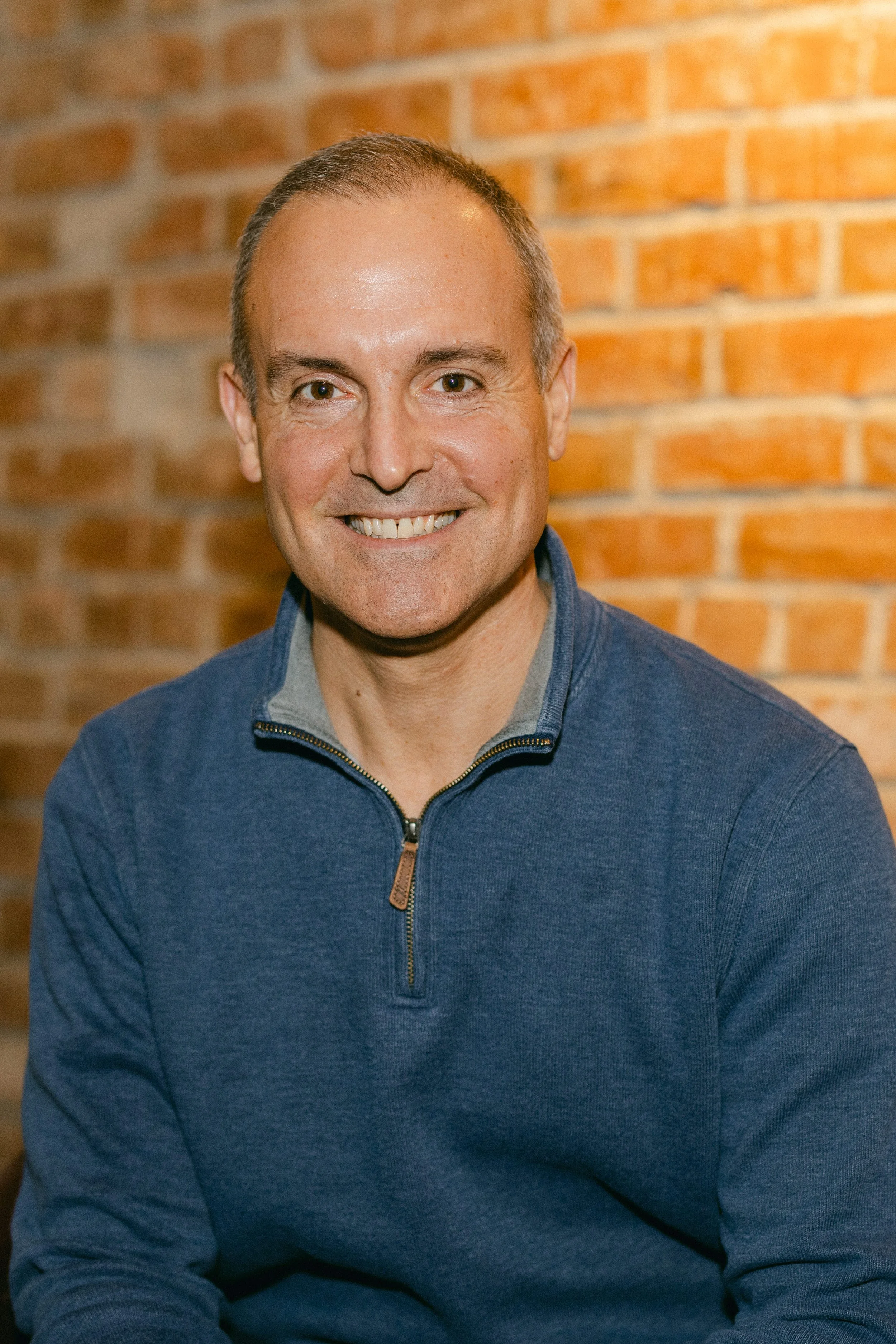 A man smiling, wearing a blue quarter-zip sweater over a gray shirt, sitting against a brick wall.