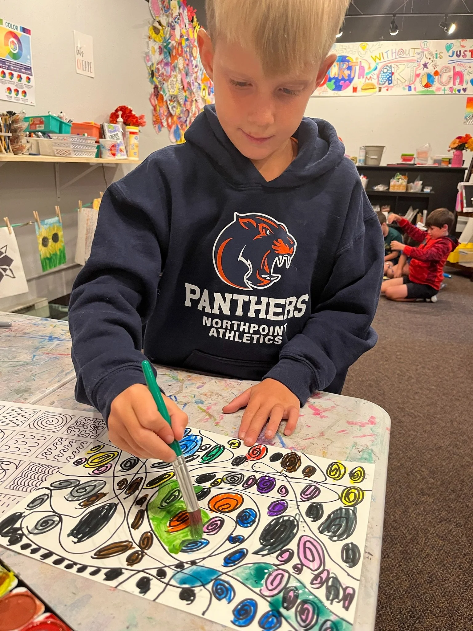 Group of young teenagers outside near a brick building, with one boy opening a box, surrounded by friends and onlookers, on a sunny day.