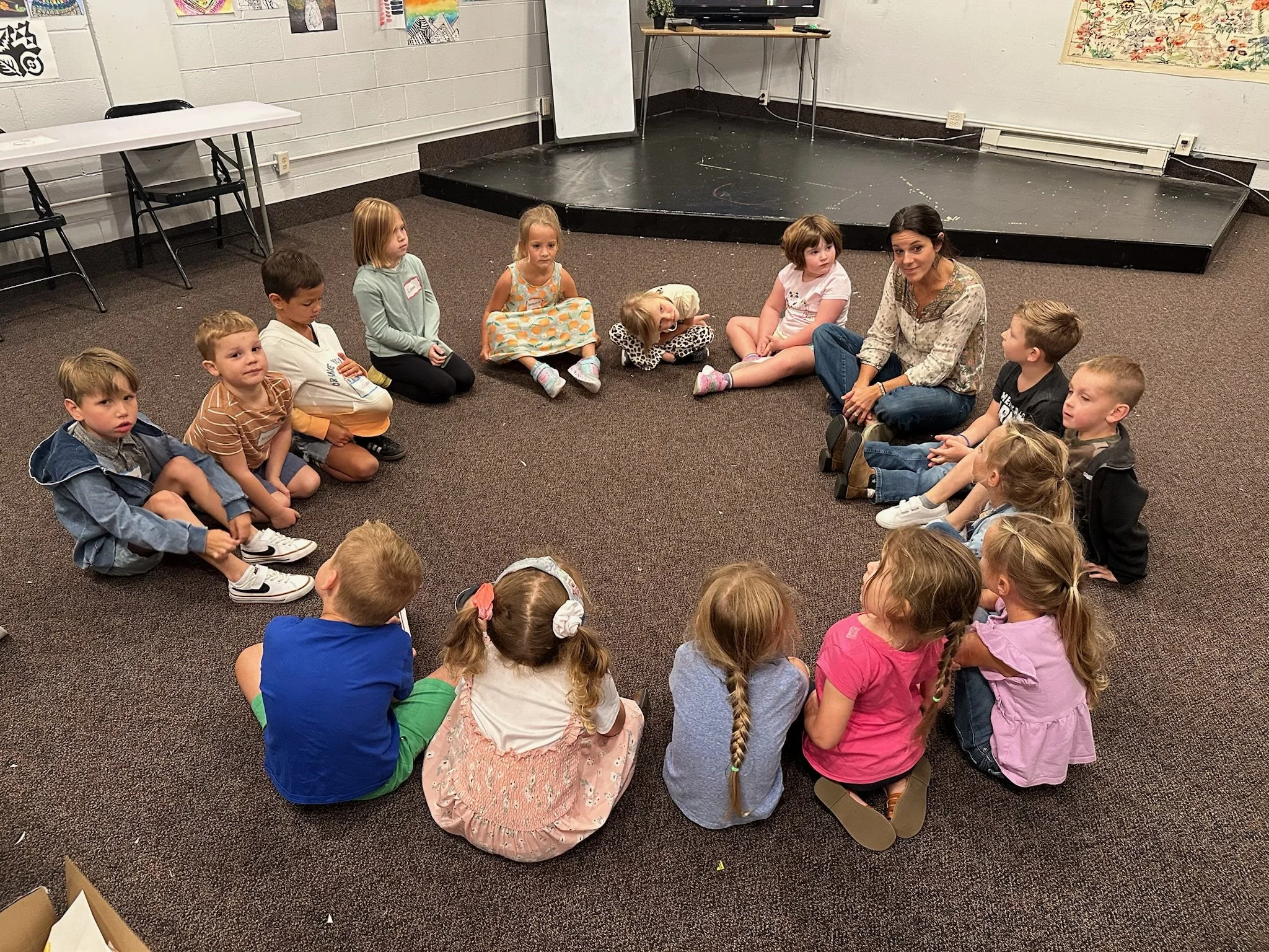 Six children sitting at two school desks with colorful bins, working on assignments in a classroom.