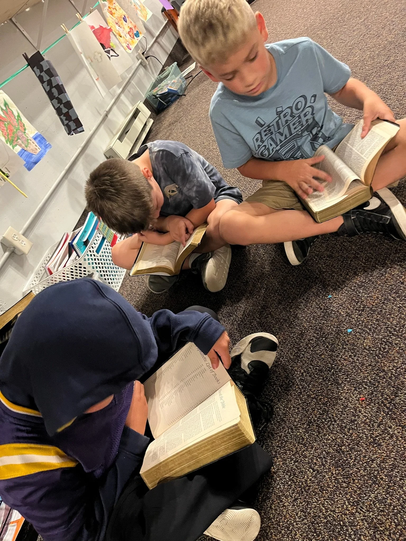 Three children sitting on the carpet playing a matching game with small white tiles, with two other children in the background reading on the floor.