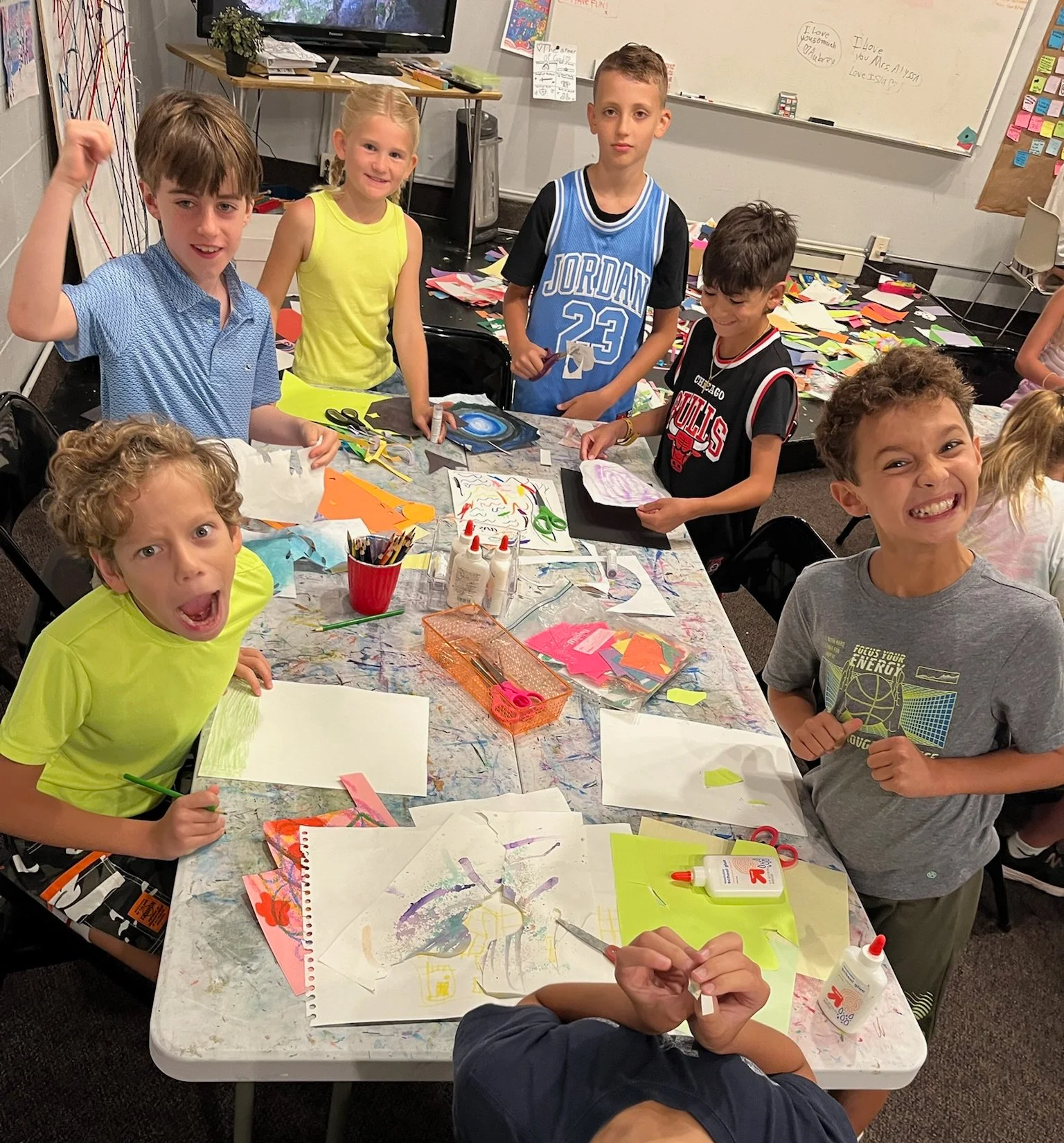 Group of fifteen teenagers standing on grass outside a brick school building, some holding boxes, smiling and posing for a photo.