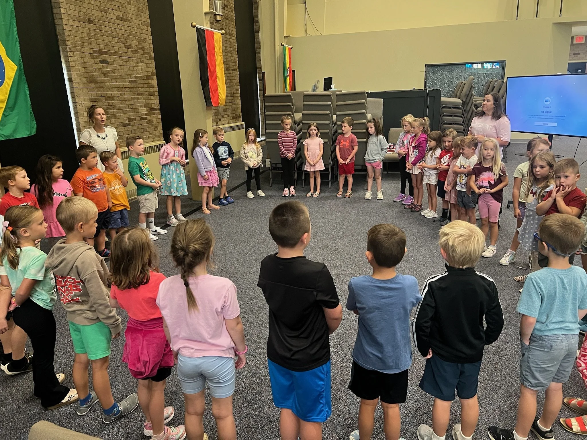 Seven smiling children standing indoors, holding drinks and backpacks, in front of a brick wall with three mounted screens.