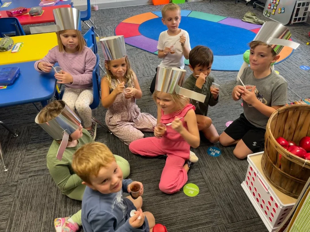 A classroom with six students seated at desks and their teacher standing at the front, smiling. The classroom has yellow walls, posters, string lights, a television, and educational materials.